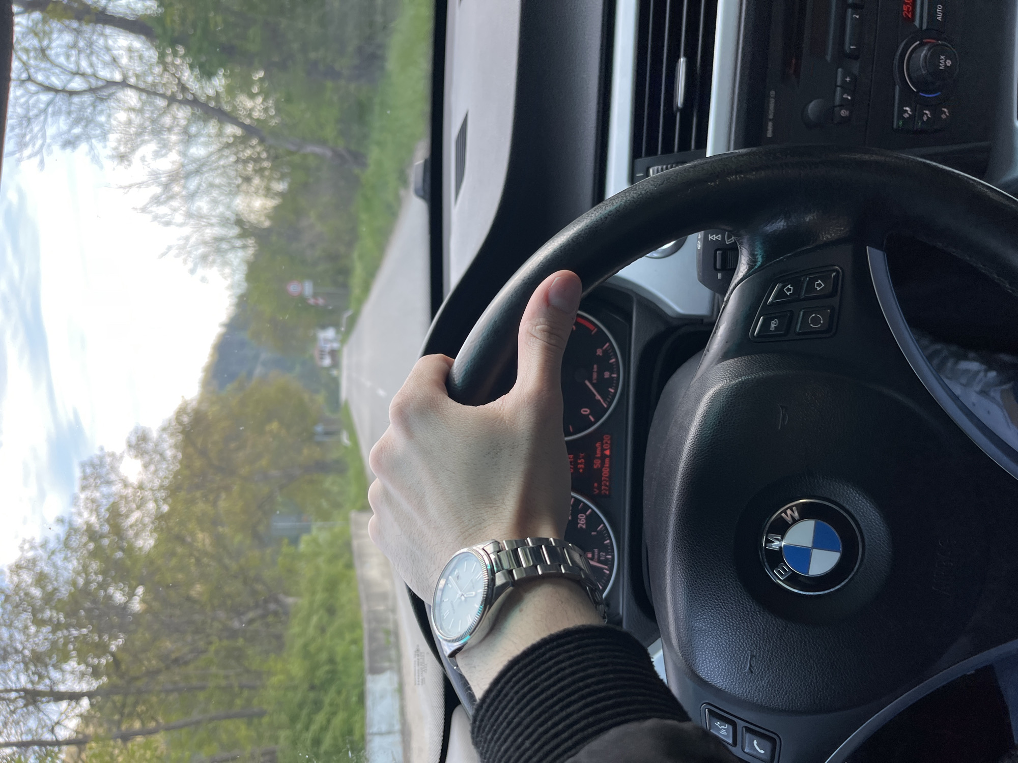 The image shows a driver's perspective from inside a BMW car. The driver's left hand is on the steering wheel, and a silver wristwatch is visible on the wrist. The dashboard and controls of the car are in focus, including the BMW logo on the steering wheel and the climate control panel. Through the windshield, a road surrounded by trees is visible. According to the car's odometer, the car has driven 272,700 km. The speedometer is at 50 km/h. Based on the scenery, the car is likely driving along a road surrounded by trees, possibly in the vicinity of Hvoina, Bulgaria.
