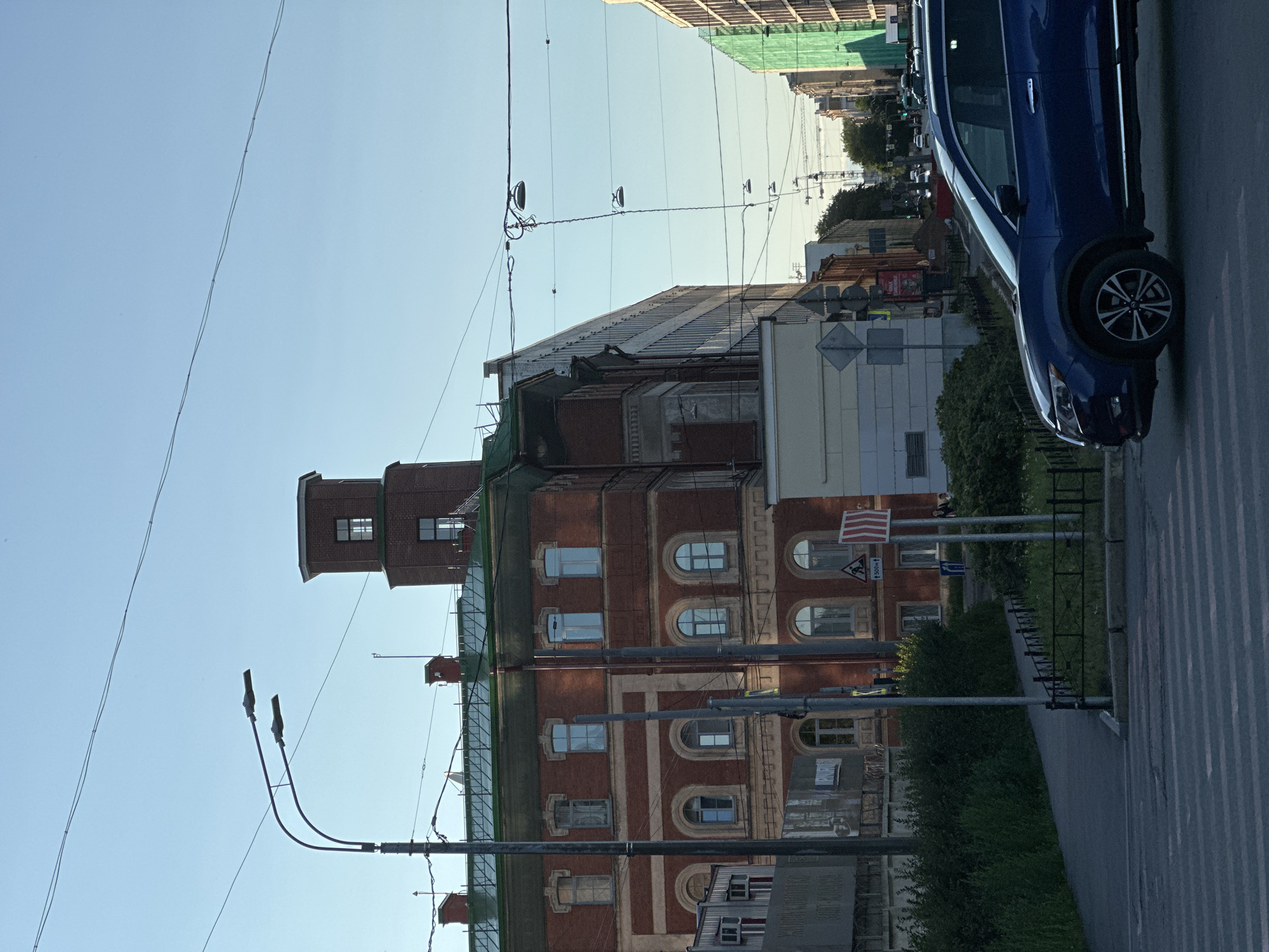 The image depicts a street scene in Saint Petersburg, Russia, likely during daylight hours with clear skies. In the foreground, a blue SUV is visible, positioned on a pedestrian crossing. The street is lined with buildings of various architectural styles. On the left, a multi-story brick building with arched windows and a green roof is prominent. To its right, a taller, modern building with a light-colored facade extends further down the street. Further down the street, on the right, another building is partially covered in green scaffolding. Overhead, numerous electrical wires and streetlights crisscross the sky, typical of urban infrastructure. A traffic light is visible in the distance. The general ambiance suggests a typical urban environment with moderate traffic and pedestrian activity.