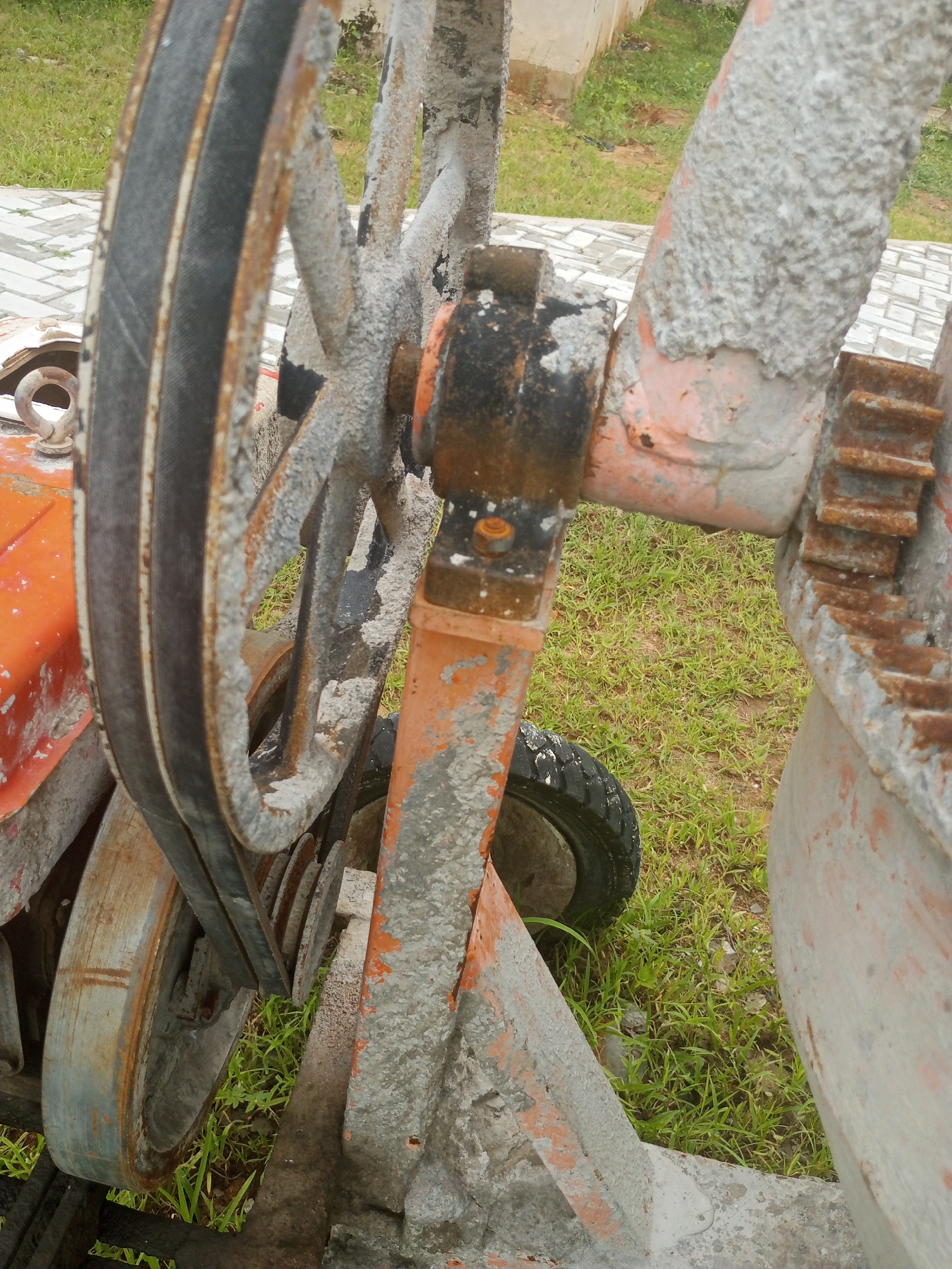 The image shows a close-up of a concrete mixer. The mixer is covered in dried concrete and shows signs of rust. A belt drive system is visible, including a large pulley and a smaller pulley. The mixer is outdoors, with grass and some paving stones visible in the background. The location is identified as Jalingo, Nigeria. There are no people or animals in the image.