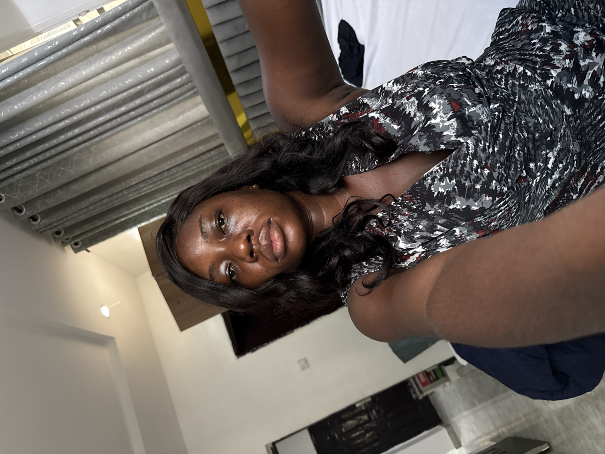 The image shows a medium-complexion woman in a printed dress, likely taking a selfie indoors. She is lying on a bed with a light-colored sheet and dark blue comforter. The room features gray patterned curtains and white walls. The specific location within the city of Iddo Tudunwada, Nigeria cannot be determined from the visual information provided.