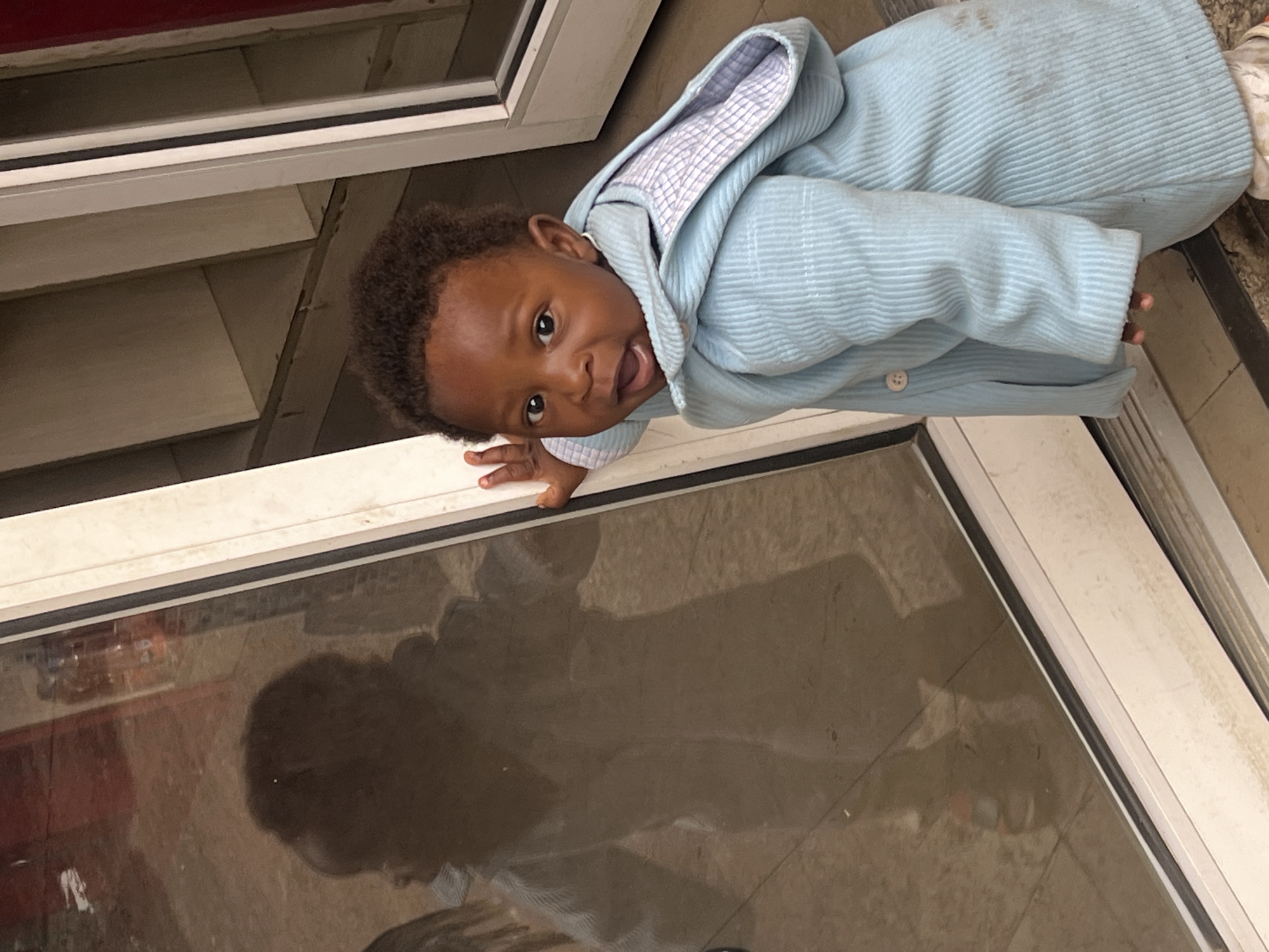 A baby with dark hair and skin, wearing a light blue hooded jacket, is leaning on the frame of a glass door or window. The door frame appears to be white. The baby is looking up and to the left with their mouth slightly open. The reflection in the glass indicates the presence of other people/objects. The setting appears to be indoors, possibly near a storefront or an enclosed area. The location is Gura Top, Nigeria.