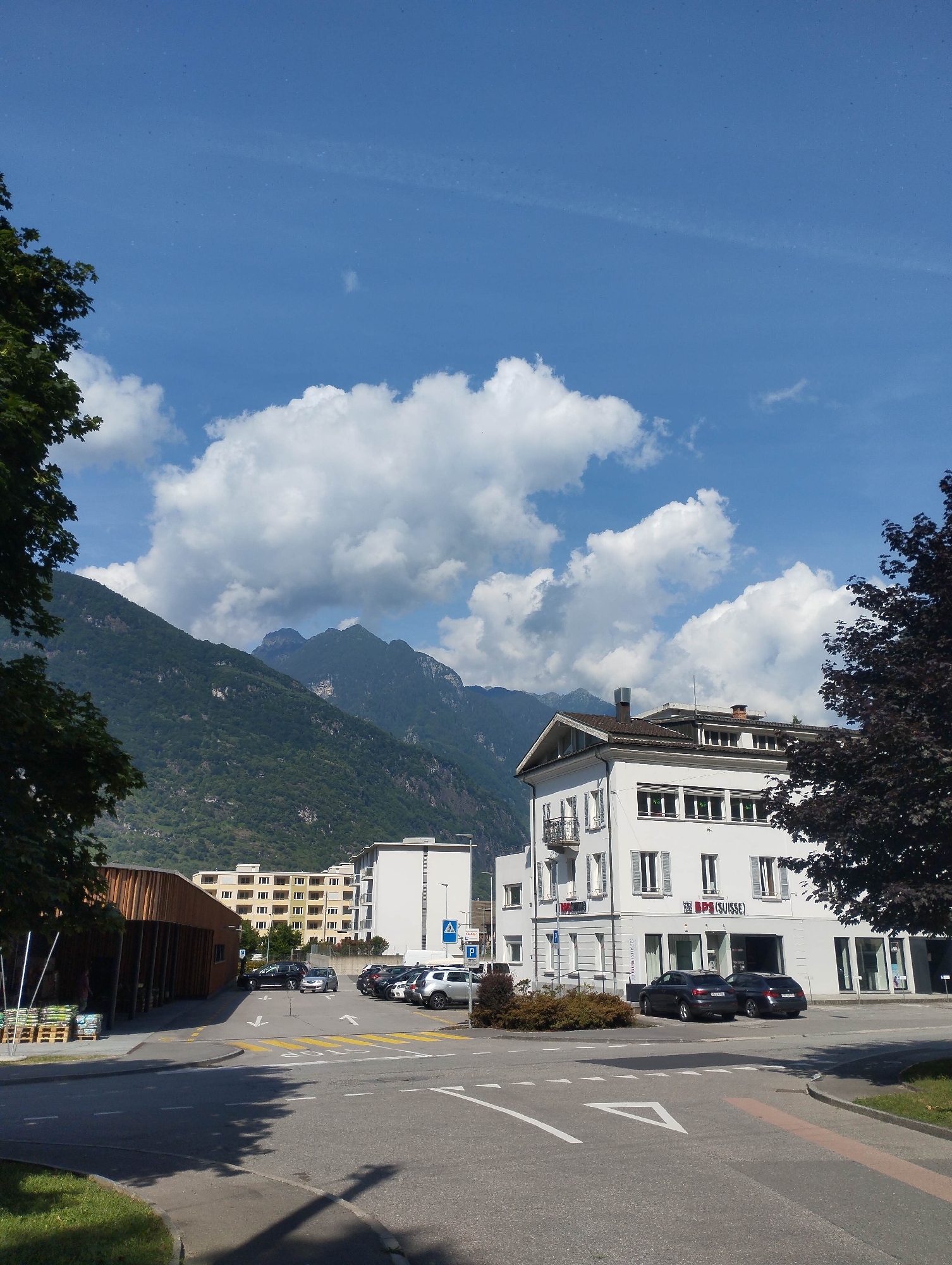 The image captures a street scene in Biasca, Switzerland, under a clear blue sky with scattered white clouds. In the foreground, a paved road with lane markings and arrows leads towards a junction. To the left, a modern building with a wooden facade and open-air shelving displaying merchandise is visible. Further down the road, a multi-story residential building with a yellow facade and numerous windows stands next to a contemporary white office building. The office building features a prominent sign reading "BPS (SUISSE)". Several cars are parked in designated areas and along the street. In the background, lush green mountains rise dramatically, partially obscured by the buildings and trees. The overall scene depicts a well-maintained urban environment in a mountainous region.