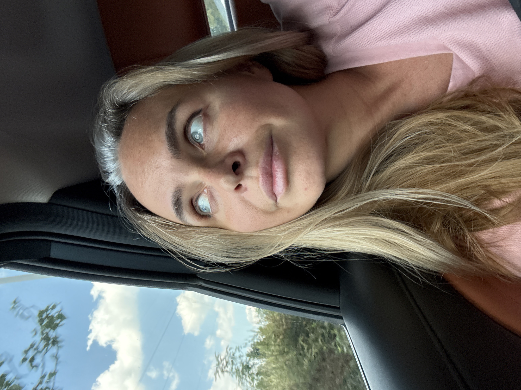 The original image shows a close-up, profile view of a middle-aged Caucasian woman looking up and to her left, out of a car window. Her blonde hair is shoulder-length, with some gray streaks visible near her temples. She is wearing a light pink, textured top. Her eyes are light blue, and her skin appears to be lightly tanned with some freckles. The background visible through the car window shows a bright blue sky with white clouds, green trees, and what appear to be utility poles. The interior of the car is visible on the right side of the frame, showing a dark gray door panel and a brown leather seat. The scene is an outdoor environment, likely a suburban or rural area, given the vegetation outside the car. There are no explicit indicators of the specific location within Wuxing District, China.