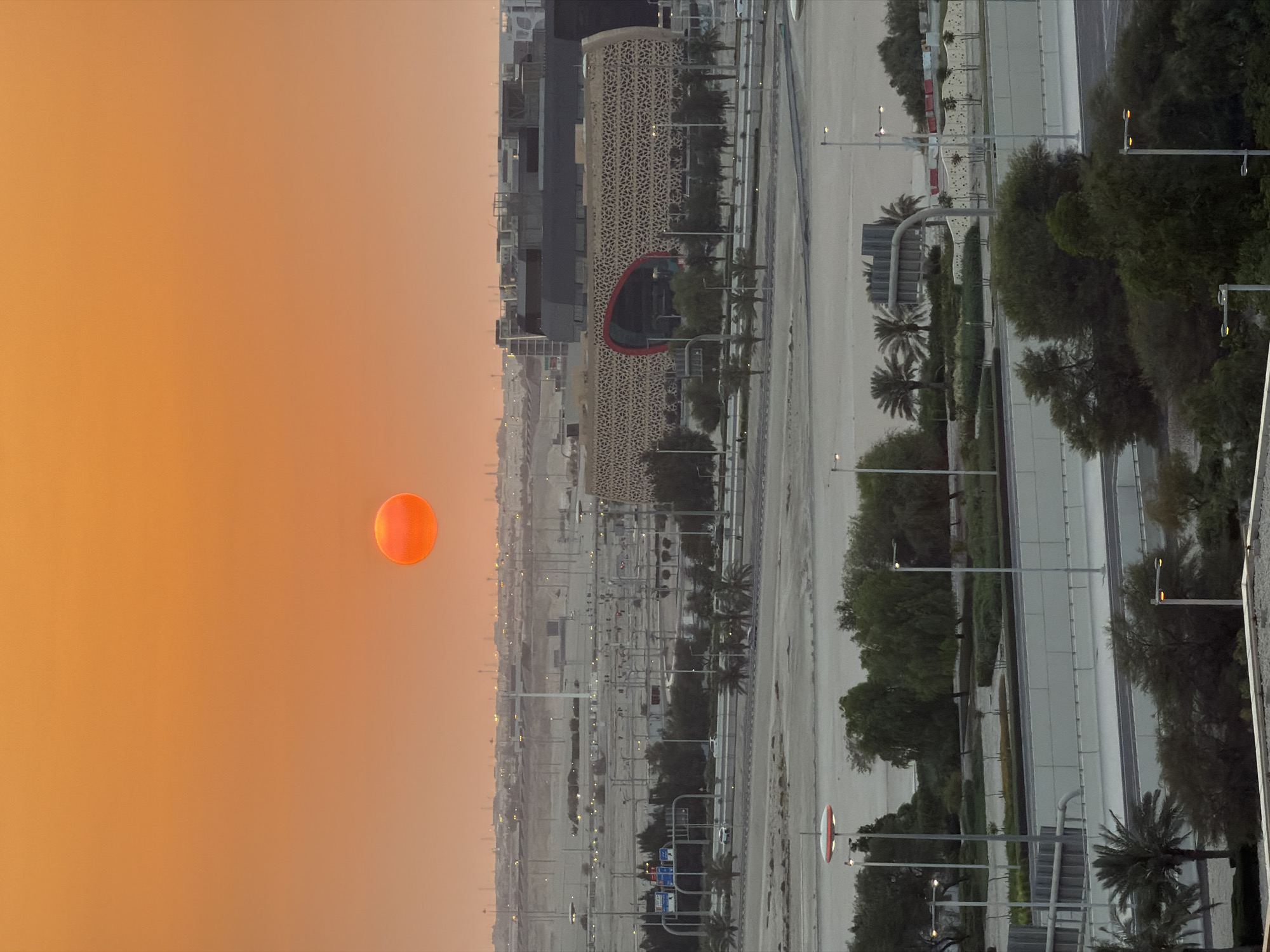 A vertical, eye-level, full shot captures a desert city skyline during sunset. The sky is a vibrant orange, with a large, circular sun hanging in the upper portion of the frame, casting a warm glow over the landscape. In the midground, a modern, curved building with an intricate, patterned facade stands out, featuring a large, arched opening. Surrounding the building are roads, streetlights, and scattered trees. The background reveals a sprawling cityscape with various structures and distant lights, indicative of urban development in a desert environment. The foreground is dominated by a multi-layered road system and lush greenery, including palm trees and other vegetation.