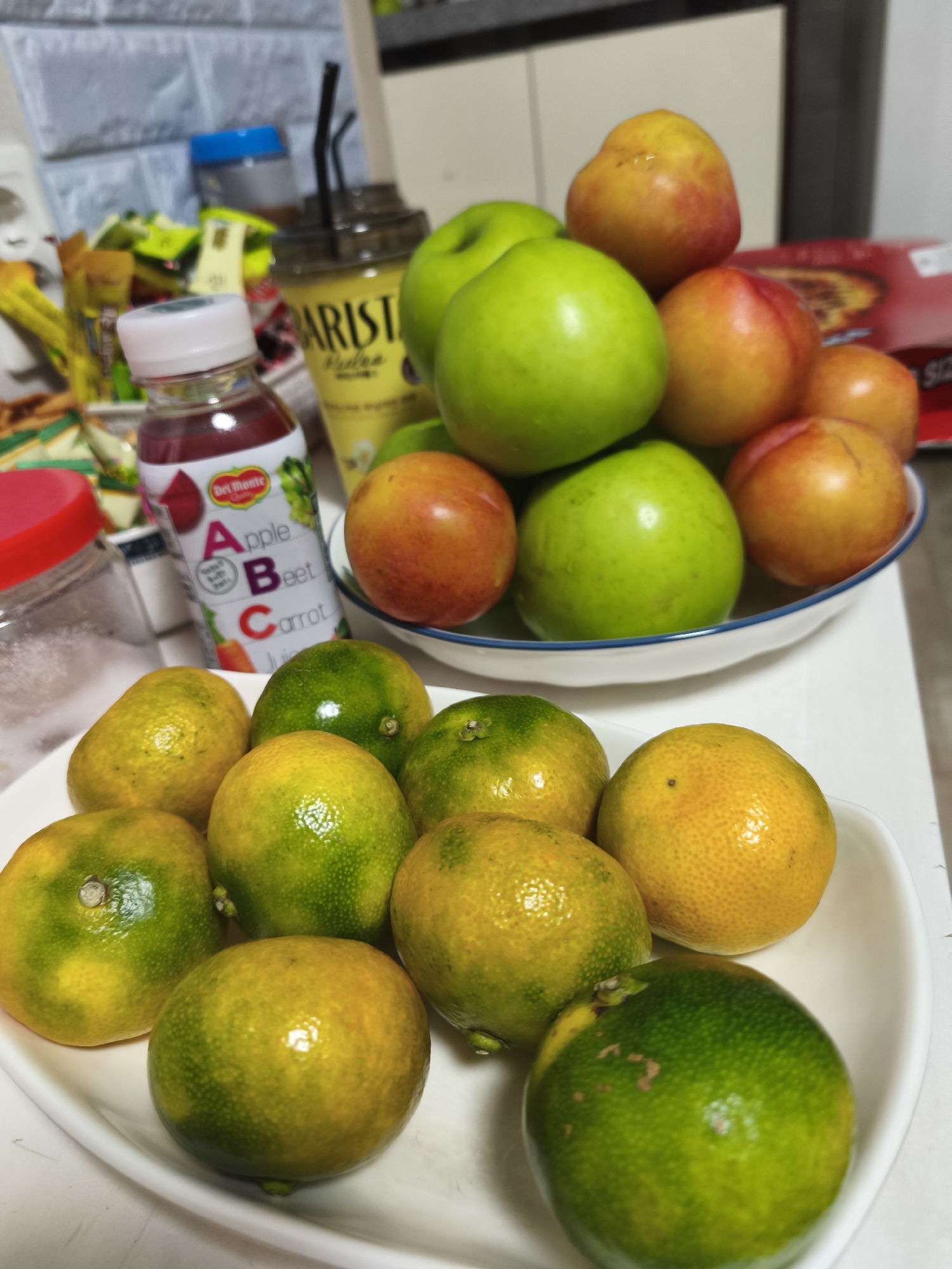 A close-up, eye-level shot captures a collection of fruits and beverages on a white table. In the foreground, a white bowl overflows with small, round citrus fruits, primarily tangerines and oranges, some exhibiting a vibrant yellow hue, while others are tinged with green. Their surfaces are textured with small pores, indicating freshness. Behind this bowl, a larger white bowl holds several green apples and reddish-yellow plums. To the left, a clear bottle with a white cap contains a deep red liquid, labeled "Del Monte Apple Beet Carrot Juice." Beside it, a clear glass jar with a red lid is partially visible. In the background, a tall, clear cup with a yellow label that reads "ARISTA" and features black straws stands prominently. Beyond that, shelves are stocked with various food items and beverages, including a blue-capped container. The backdrop appears to be a tiled wall, possibly in a kitchen or pantry. The lighting is bright, suggesting an indoor setting. The overall scene is one of an organized display of fresh produce and drinks, likely in a residential environment within the city of Asan-si, South Korea, given the context.