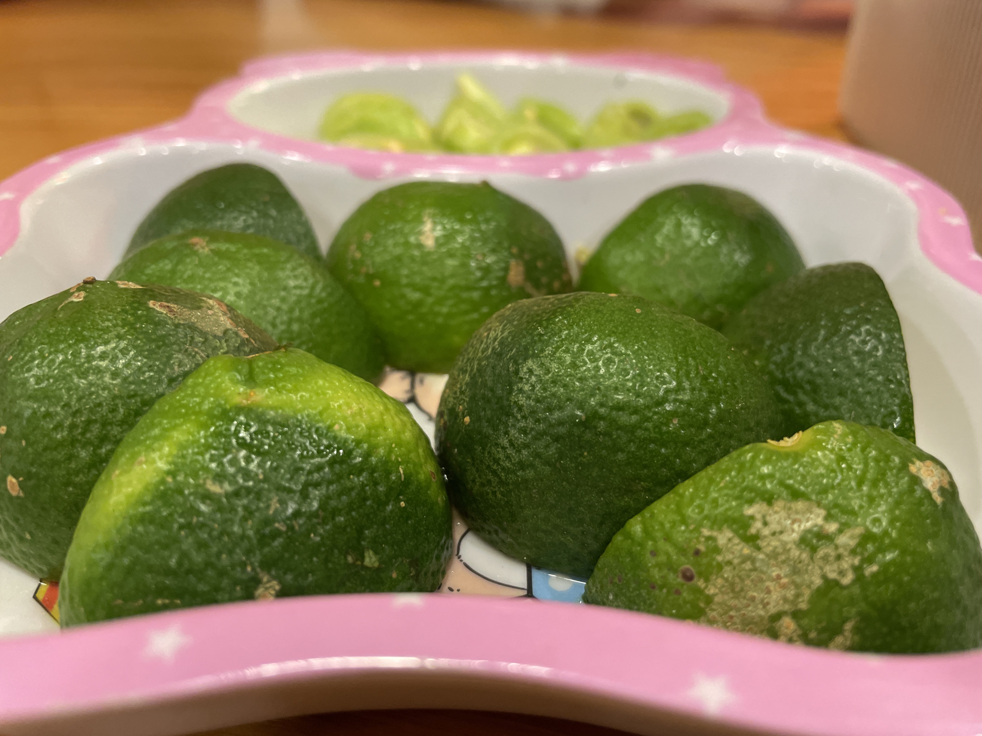 The image shows a close-up of several green limes arranged in a pink-rimmed dish. The dish has a slightly heart-shaped or scalloped edge with white and pink detailing. The limes are mostly round with a textured rind, and some have light brown or tan blemishes. In the background, out of focus, there appears to be a light green substance in a section of the dish, possibly another food item. The lighting suggests an indoor setting, possibly a kitchen or dining area, with a warm-toned wooden surface visible in the far background. The focus is sharply on the limes in the foreground, with the rest of the scene progressively blurred. There is no visible text in the image. The overall impression is of fresh produce prepared for consumption or presentation.