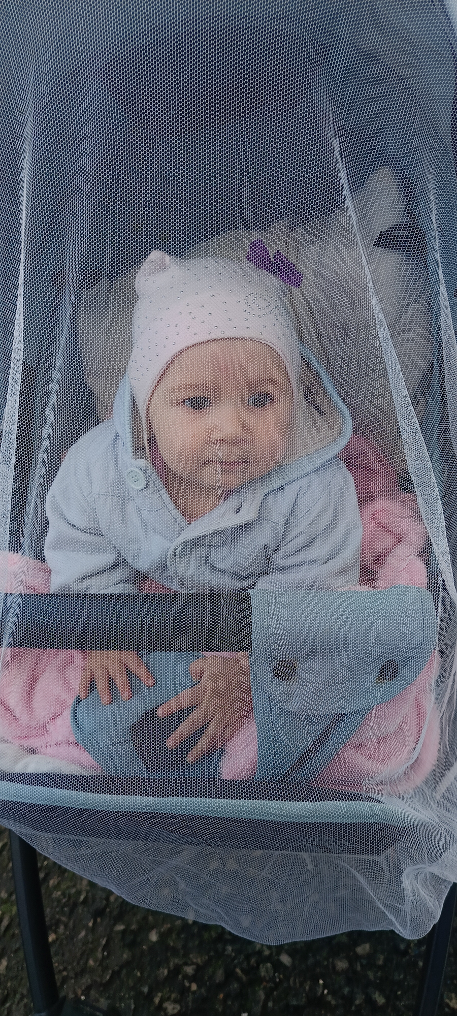 An infant, wearing a light pink hat adorned with rhinestones and a small purple bow, is nestled in a stroller covered by a white mesh net. The baby is dressed in a light blue hooded jacket with a single visible button. Their hands, one resting on a pink blanket and the other near a blue cushion, are visible through the net. The stroller appears to be outdoors, with a dark, textured surface resembling pavement or soil visible at the bottom of the frame. The scene is likely located in or around the city of Khokhly, Russia.
