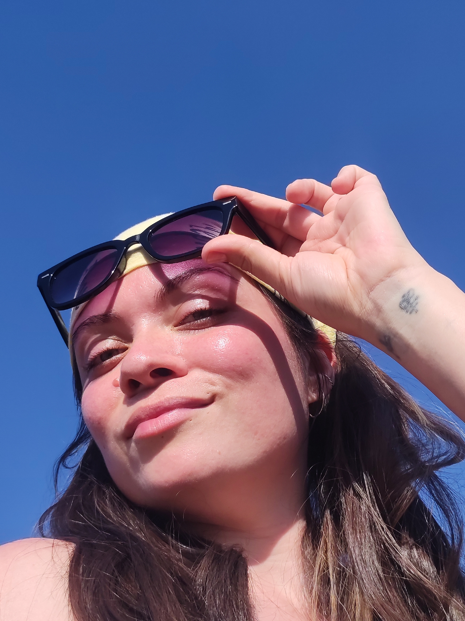 A low-angle, medium shot depicts a fair-skinned woman against a clear blue sky. She wears a yellow headband and black-framed sunglasses, which she is lifting slightly with her right hand. Her makeup includes pink eyeshadow and tinted lip gloss. A small, dark tattoo is visible on her wrist. Her long, dark brown hair falls to the side. The location is not discernible.