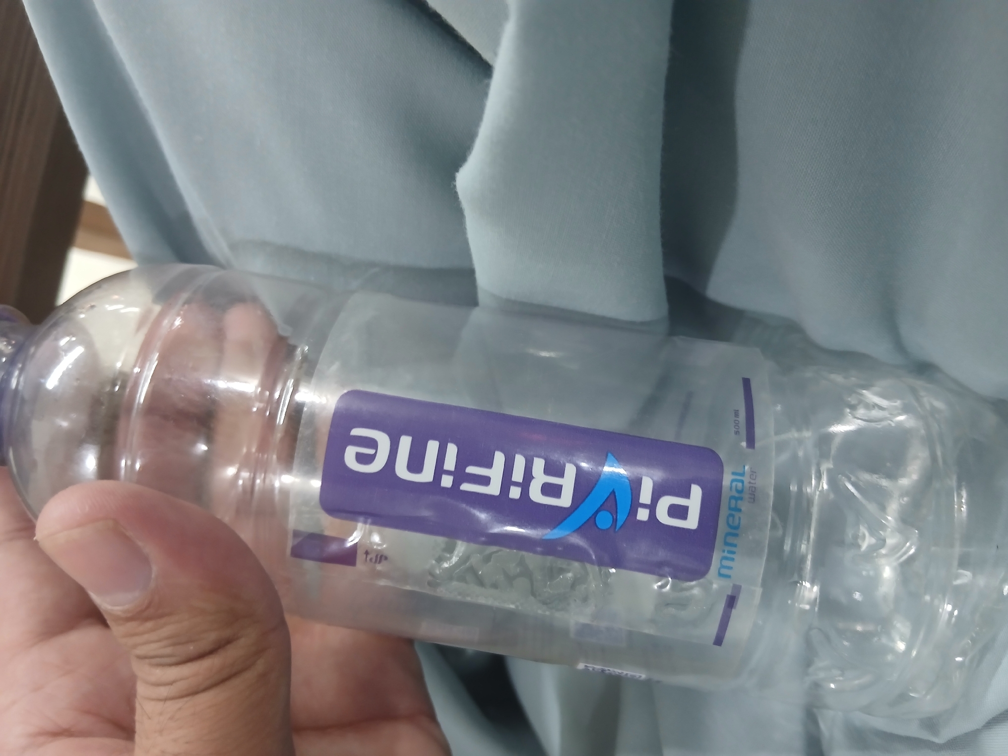 A hand is holding a clear plastic water bottle labeled "Pi RìFine" and "MINERAl water". The bottle is mostly full of water and has a purple label with white text. The background is a light blue fabric, possibly clothing or a blanket. The scene is indoors, with hints of wooden furniture in the upper right corner. The location of Peshawar City Tehsil, Pakistan is not discernible from the image.