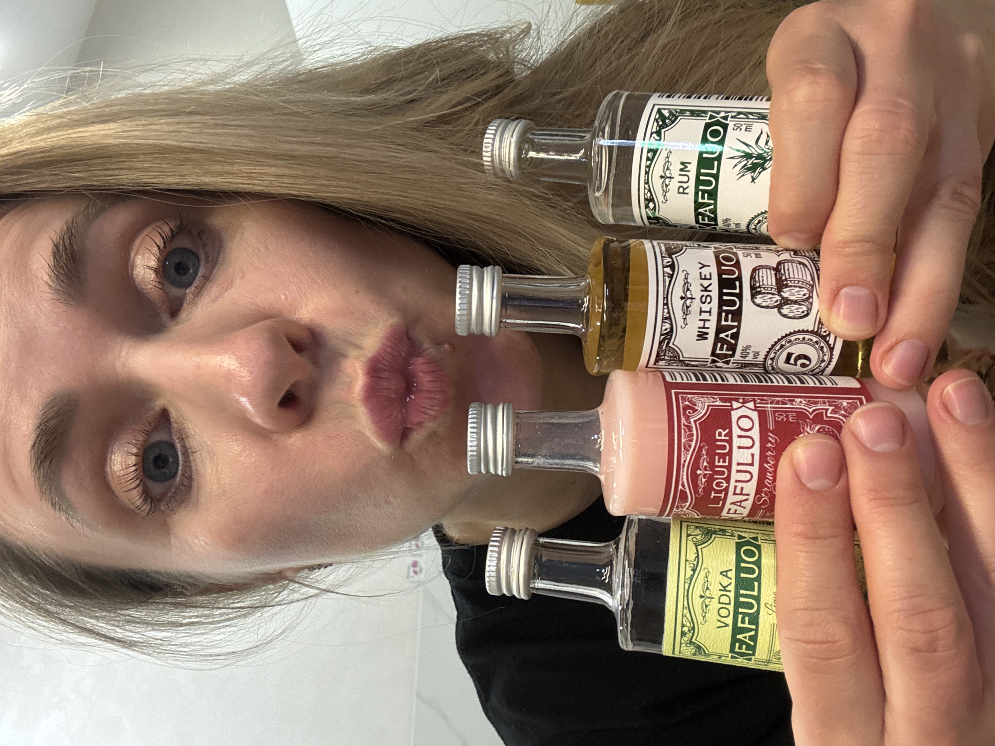 A close-up, rotated portrait-style image features a person, likely a woman, with long blonde hair partially covering the upper part of her head. Her right blue eye is prominently visible, looking towards the camera, along with a portion of her nose and lightly made-up, puckered lips. She holds four miniature alcohol bottles in her left hand, positioned in the lower right section of the frame.
The bottles are labeled with the brand "FAFULUO" and display various spirits. From top to bottom, the labels clearly show: "RUM FAFULUO" with "50 ml" and "40% vol" indicated, featuring a plant illustration; "WHISKEY FAFULUO" also at "40% vol," showing barrels and the number "5"; "LIQUEUR FAFULUO Strawberry"; and "VODKA FAFULUO Limo" (likely "Lemon" or "Lime"). The person's fingers grip the bottles, showcasing neatly manicured nails. The background is a plain, light surface, suggesting an indoor setting. The puckered lips convey a playful or affectionate emotion. The location context for this media file is Chaikovsky, Russia.