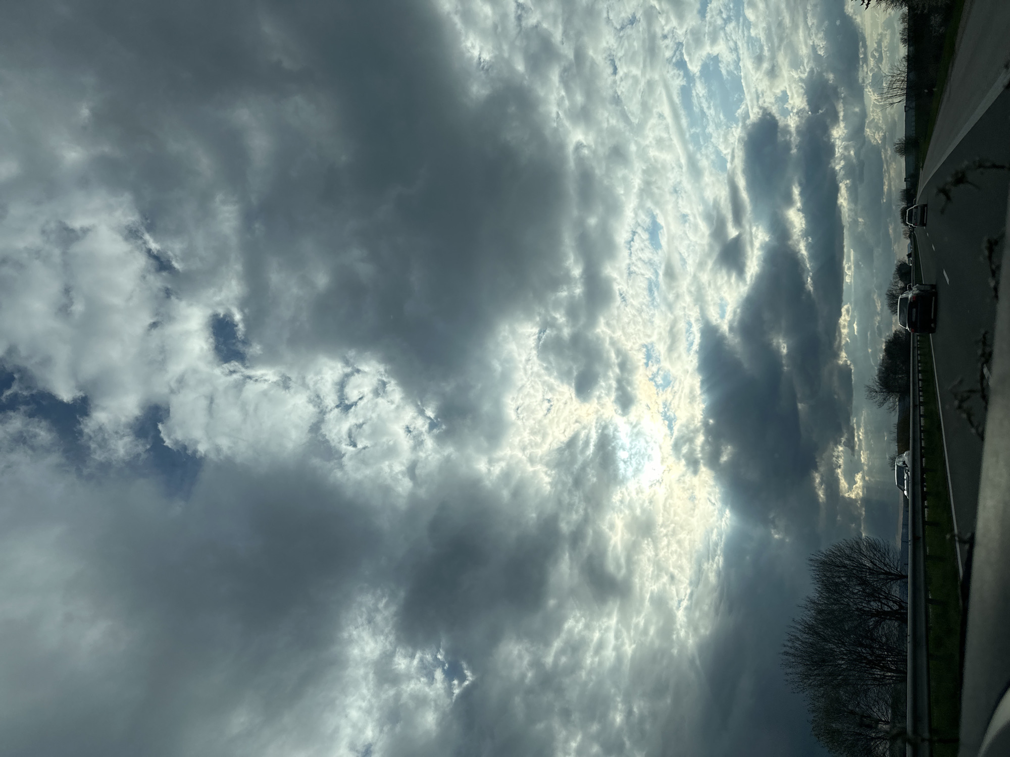 The image captures a view from a moving vehicle, likely on a highway, with the primary focus being the sky. The sky is filled with a mix of dark, heavy clouds and patches of brighter, sunlit areas. The light is breaking through the clouds in places, creating a dramatic effect. The highway is visible on the right side of the frame, with a guardrail, some trees, and several cars in motion. The scene does not indicate that the location is in the city of Gabarnitsa, Bulgaria.