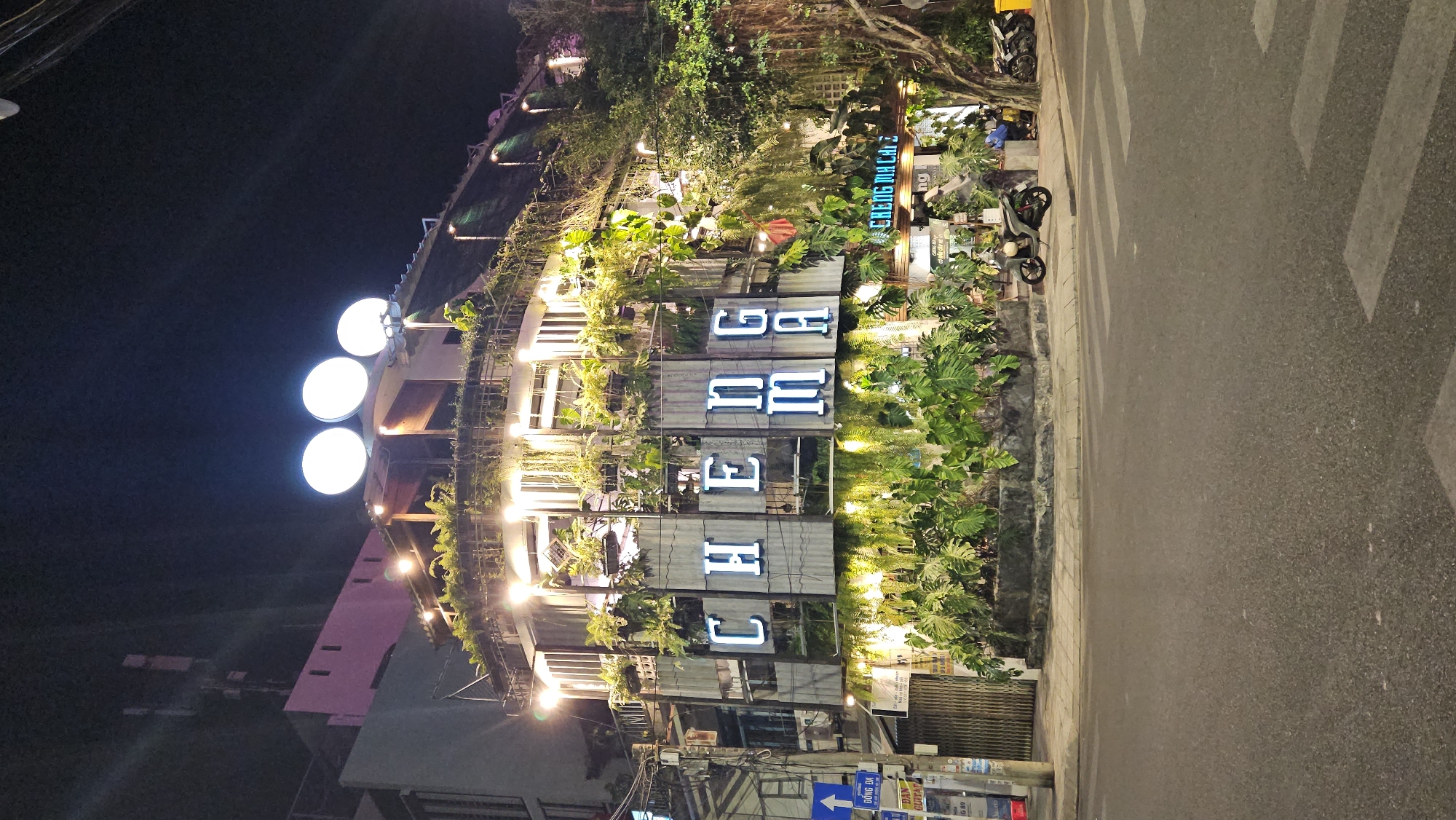 At night, an upward-angled shot captures a multi-story building in Nha Trang, Vietnam. The building's façade is adorned with lush greenery, partially obscuring neon-lit block lettering that reads "CHE BA FE." Adjacent to the building, a sidewalk and a section of street are visible, featuring a crosswalk. A parked motorcycle is nearby. Above the building, three large, round lights illuminate the scene against the dark sky. The structure's architecture includes multiple levels with lit windows and balconies. Other buildings of varied heights and colors are visible in the surrounding area. Utility poles and wires are present. Street signs appear in the foreground.
