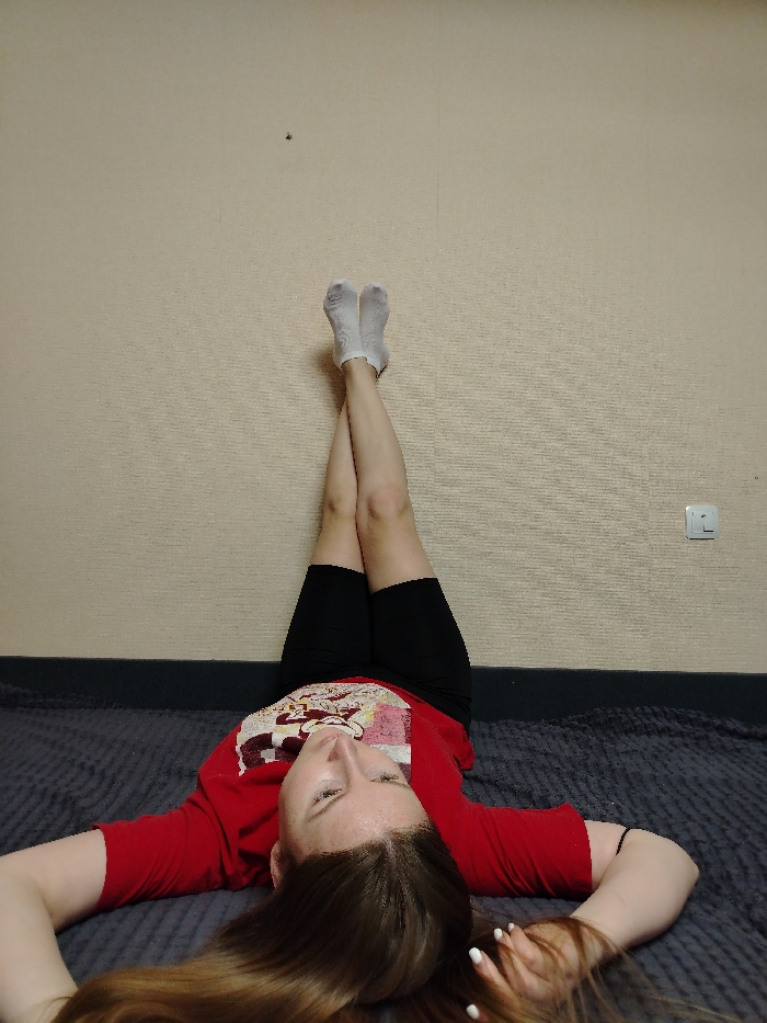 In an indoor setting in Chaikovsky, Russia, a single individual is depicted lying supine on a dark gray textured blanket. The person has light brown hair and is wearing a red short-sleeved t-shirt with a graphic design, black shorts, and white ankle socks. Their legs are elevated vertically against a light beige textured wall, crossed at the ankles. Both arms are extended laterally, resting on the blanket. A white electrical outlet with two circular sockets and a switch is visible on the wall to the right of the frame, and a small dark blemish is present on the upper-left portion of the wall.