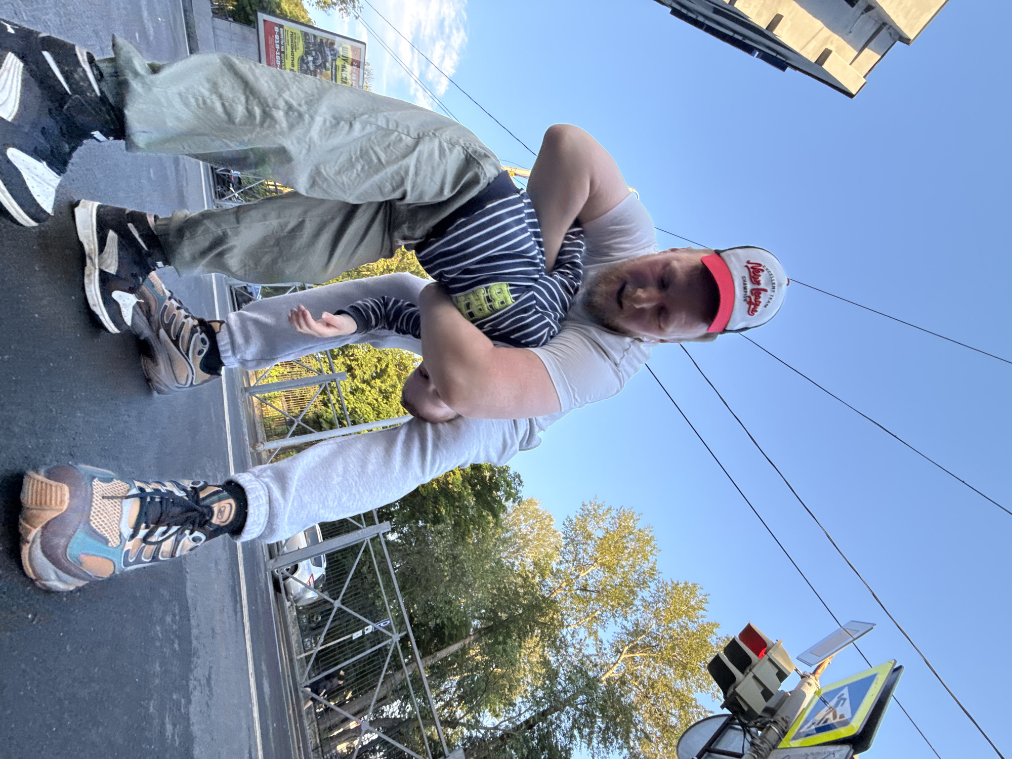 The image depicts two individuals in a low-angle shot on a city street, possibly in Perm, Russia, under a clear blue sky. A tall, modern building with a light-colored facade is visible on the left, and power lines stretch across the sky. On the right, a traffic light showing a red signal and pedestrian crossing signs are mounted on a pole. Lush green trees line the background, with a metal fence separating the street from a sidewalk or pathway. A billboard with Russian text and imagery is also present on the left side of the frame.
The two individuals appear to be a man and a child engaged in a playful interaction. The man, wearing a white t-shirt, light grey sweatpants, and a red and white baseball cap with the text "EXCELLENT TEAM New league CHAMPION," is holding the child in a wrestling-like pose. The child is wearing a striped long-sleeved shirt and light grey sweatpants. Both are wearing athletic footwear. The man is on the right, and the child is to his left, partially obscured by the man's arms. The ground is asphalt, indicating a street or road.