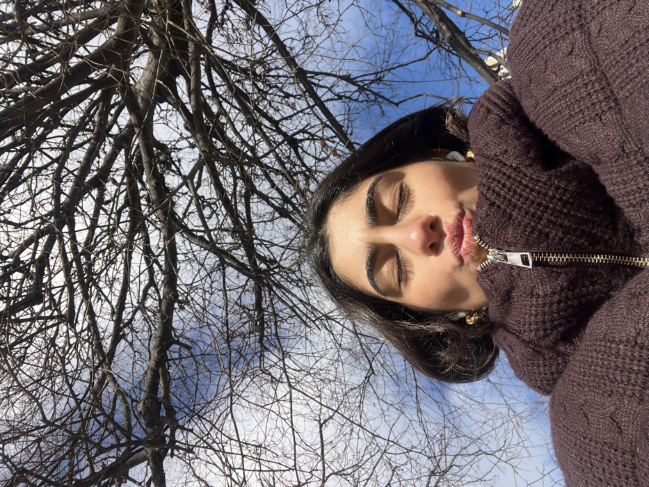 A female subject is depicted in an upward-angled close-up shot, positioned against a clear blue sky with some scattered white clouds and a dense network of bare tree branches, characteristic of deciduous trees in dormancy. The subject has dark hair and her eyes are closed. Her lips are pursed forward in a "kissing" pose. She wears white wireless in-ear earbuds and small, ornate gold-colored earrings. Her upper body is clad in a dark brown, textured, possibly knitted, high-collared jacket with a visible silver-toned zipper pull at the front. The lighting suggests direct sunlight. This scene is situated outdoors in Paris, France.