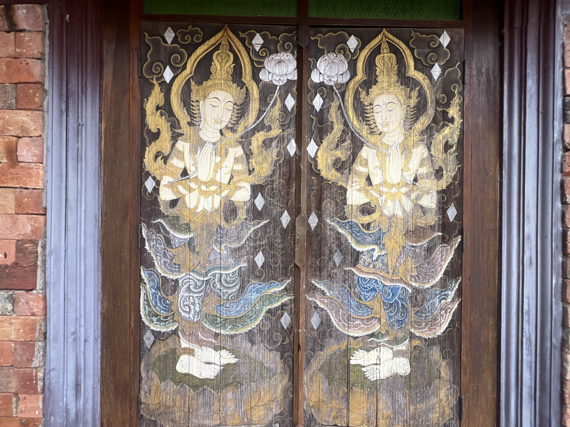 The image shows a pair of wooden doors with traditional Thai paintings of two divine beings, possibly angels or deities, on each door. The figures are depicted with golden headdresses, ornate clothing in white, blue, and gold, and are shown with their hands pressed together in a gesture of prayer or greeting. They are standing on what appears to be lotus flowers, which are also painted in muted tones. The background of the paintings is dark wood, with swirling golden patterns and diamond-shaped motifs. The doors are set within a brick wall on the left and a dark wooden frame on the right. The overall aesthetic suggests a religious or cultural setting.
