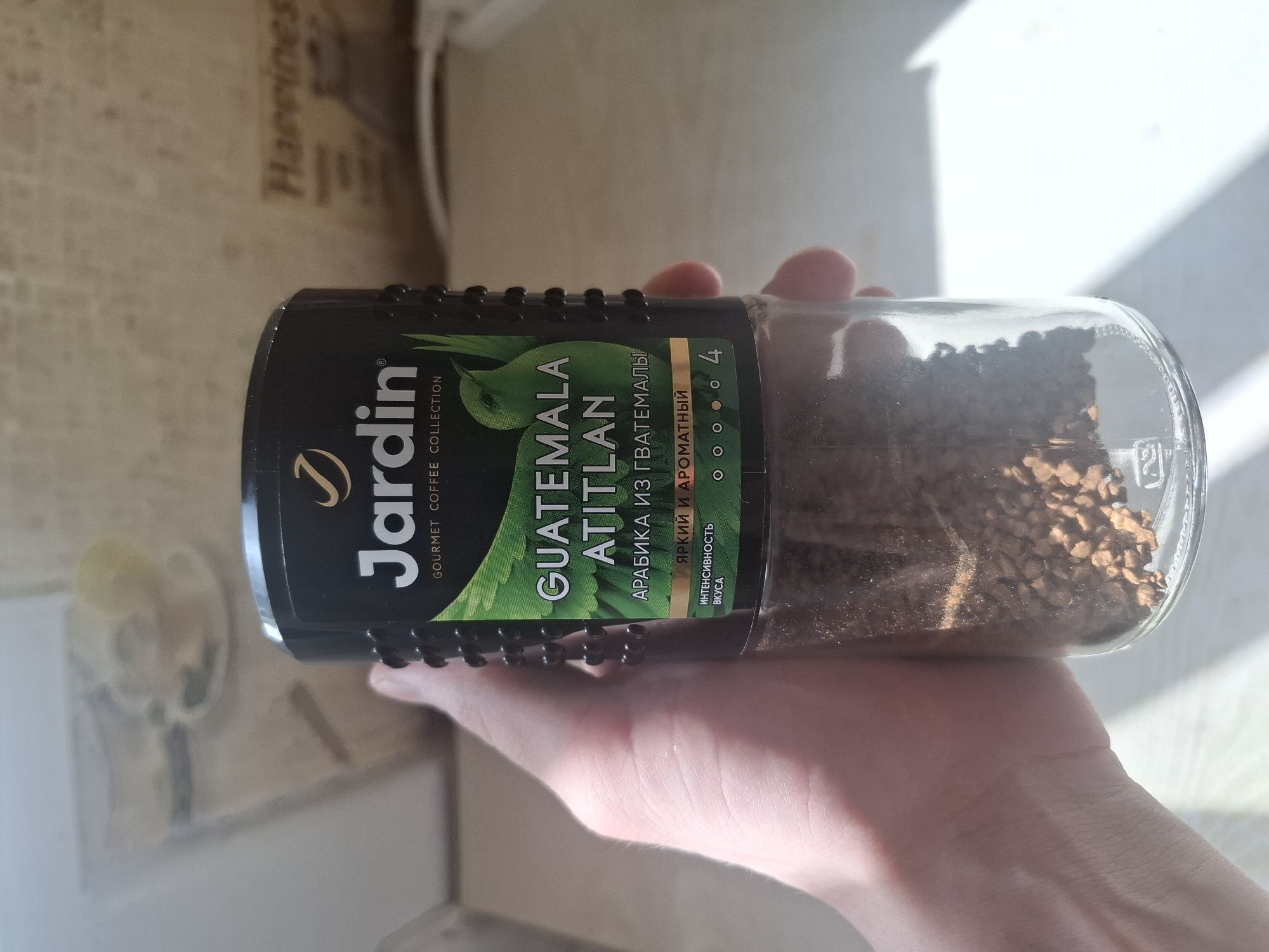 A person's hand holds a cylindrical glass jar of instant coffee horizontally against a brightly lit, blurry background. The jar, about half full of dark brown granular coffee, features a prominent black label. The label displays the brand "Jardin" and "GOURMET COFFEE COLLECTION." The coffee type is specified as "GUATEMALA ATITLAN," with accompanying Cyrillic text "АРАБИКА ИЗ ГВАТЕМАЛЫ" (Arabica from Guatemala). Further text on the label reads "ЯРКИЙ И АРОМАТНЫЙ" (Bright and Aromatic) and "ИНТЕНСИВНОСТЬ ВКУСА" (Flavor Intensity), visually represented by four out of five filled circles. The hand holding the jar is light-skinned, with visible veins and skin texture. The background is indistinct, showing light-colored surfaces and strong sunlight casting shadows. Faintly visible in the upper right corner of the background is the word "Happines." The natural light and shadows suggest it is daytime. The setting appears to be an indoor environment, likely a home, with no specific activity beyond holding the coffee jar.