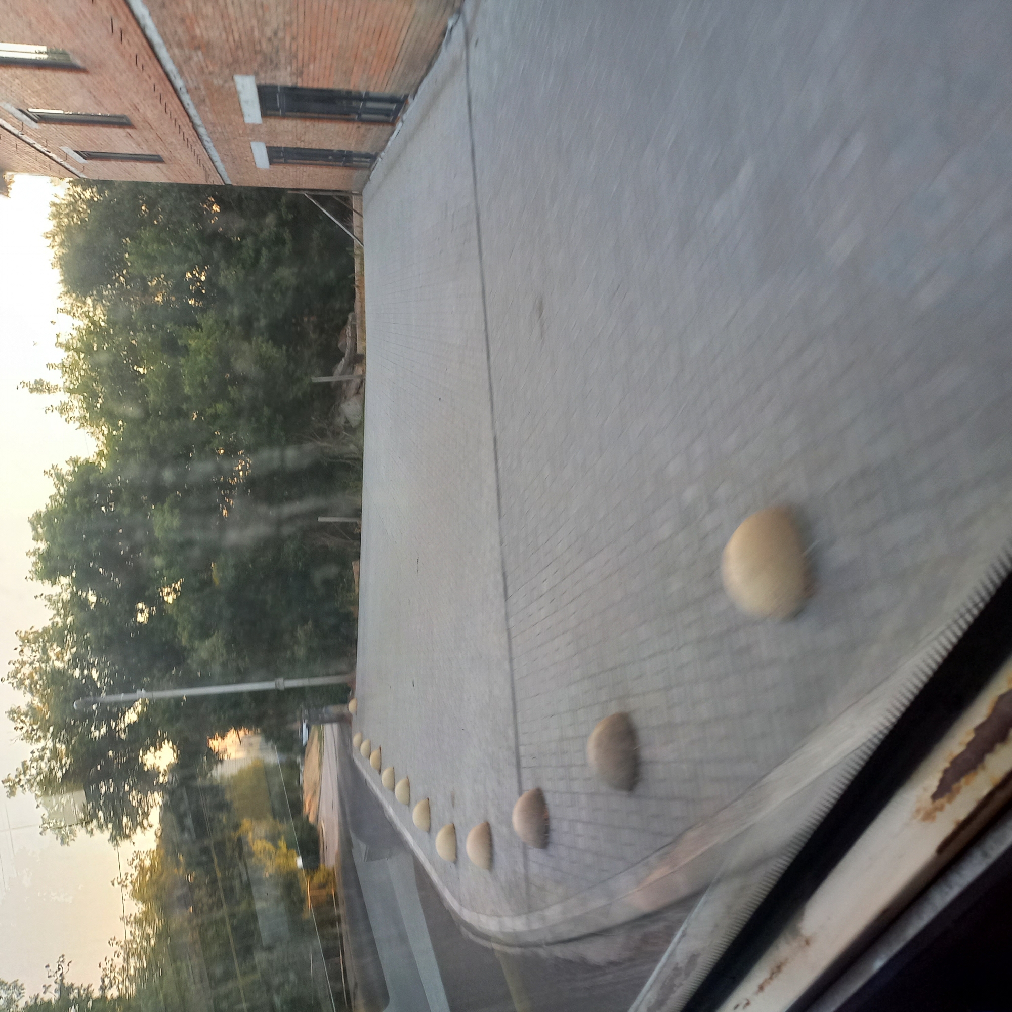 The image shows a cobblestone pavement with a curved border lined with yellow, dome-shaped bollards. A brick building with windows and doors is visible on the right side of the frame. Trees are in the background, and the sky appears overcast or hazy, suggesting it might be early morning or late evening. The scene is outdoors, likely in an urban or semi-urban setting. The perspective suggests the photo was taken from inside a vehicle, looking out through a window, as evidenced by the reflections and the distressed frame of the window in the foreground. The location is not specified in the image.