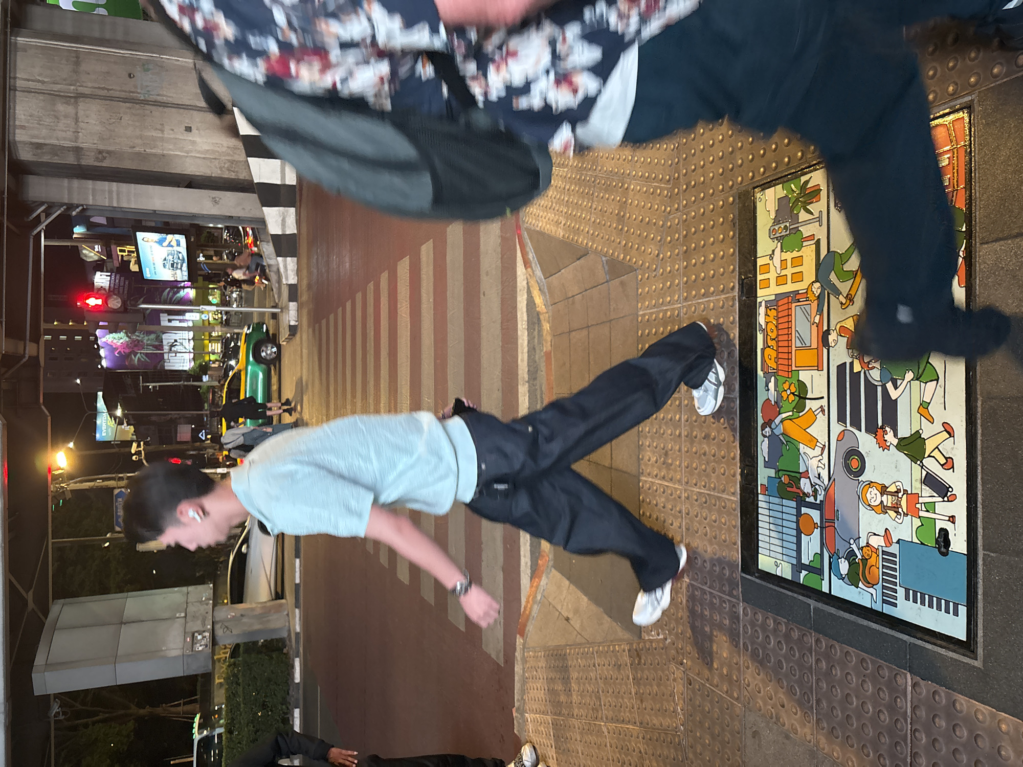 The image shows a street scene, likely at night, in what is presumed to be Bangkok, Thailand. The focal point is a painted or mosaic artwork embedded in the sidewalk, depicting cartoon-style people and vehicles, typical of city life. Two pedestrians are in the frame: one appears to be walking across the crosswalk and has a light green shirt and dark pants. The other person in the upper right with dark clothing and a floral pattern shirt is mostly cut off from the frame. A crosswalk is visible, marked with horizontal red and white stripes. Beyond the sidewalk, there's a street with cars, including a yellow taxi, and buildings with signs. A pedestrian is shown wearing earbuds and checking their wrist watch. The setting has the feeling of a commercial district with stores and traffic.