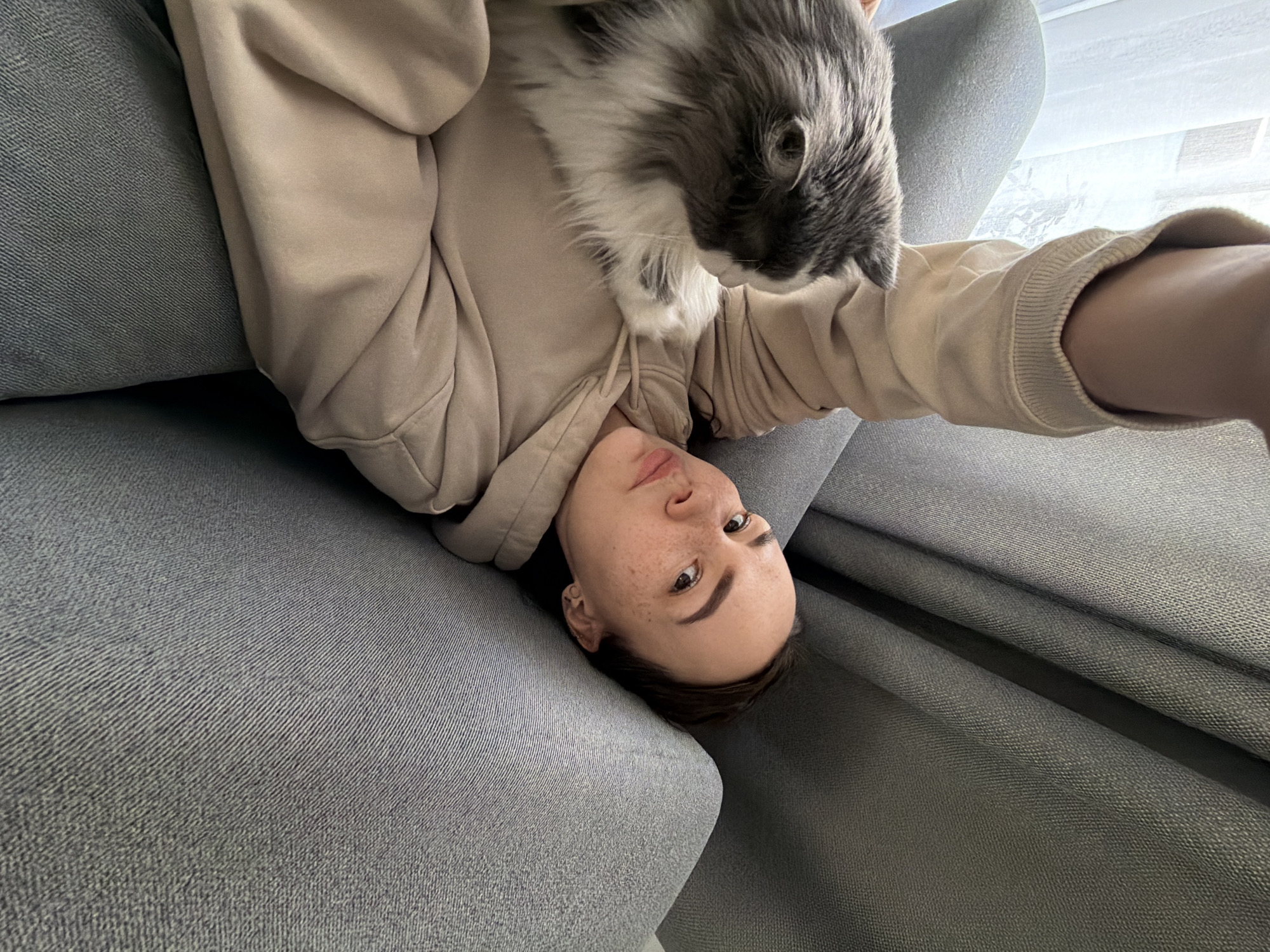 An intimate, high-angle selfie captures a young person and a fluffy gray and white cat in an indoor setting in Chaikovsky, Russia. The person is lying on their back on a textured gray sofa, looking upwards towards the camera with a neutral, slightly relaxed expression. They wear a light-colored, possibly beige or tan, hooded sweatshirt. Notable facial features include visible freckles and dark hair. The cat is positioned on the person's upper chest or shoulder, its head turned downwards, observing the scene. In the background, daylight filters through a window with blinds, indicating a daytime setting. The image portrays a quiet, casual moment between a pet owner and their cat. There is no visible text.