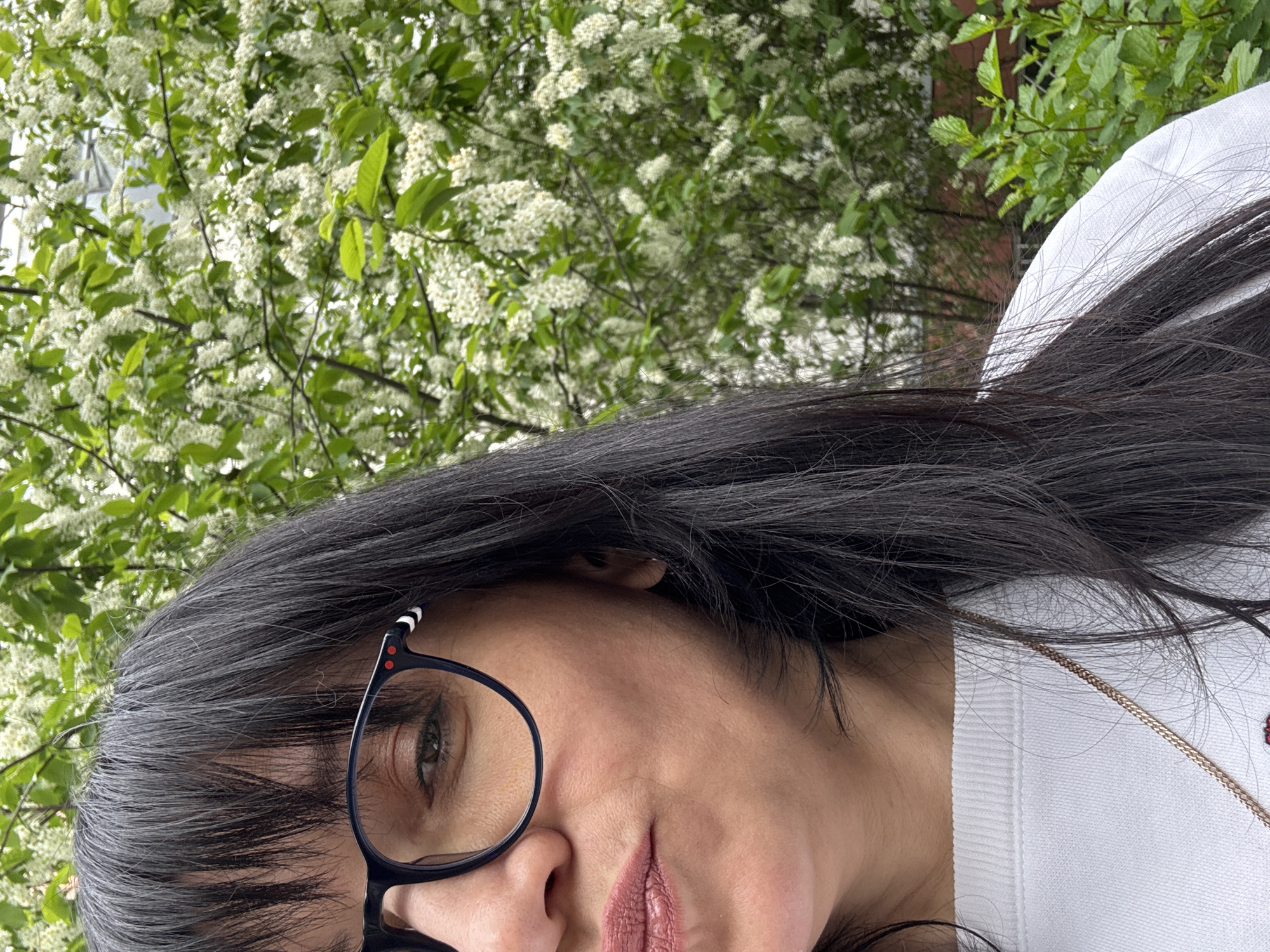 A woman with dark hair and glasses is partially visible in the foreground. She is wearing a white top and a delicate gold necklace. The background consists of lush greenery, including a flowering tree or bush with white blossoms. The exact location in the city of Chaikovsky, Russia cannot be determined from the image.