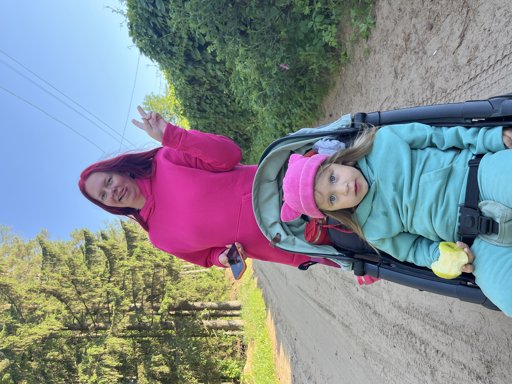 The image captures an adult woman and a young child outdoors on a bright, sunny day in what appears to be Chaikovsky, Russia. The photograph is oriented vertically, rotated 90 degrees clockwise.
The woman, standing on the left side, has long reddish-brown hair and is dressed in a vibrant pink hooded sweatshirt and matching sweatpants. She has a nose piercing and is making a peace sign with her right hand, while holding a smartphone in her left. Her expression is cheerful, with a slight smile and her tongue playfully out.
To the right, a young child with blonde hair and clear blue eyes is seated in a stroller. The child is wearing a light teal hooded sweatshirt and matching pants, along with a pink hat featuring ear-like embellishments. Secured by a harness, the child holds a partially eaten green apple in their right hand and looks directly at the camera with an attentive gaze.
The setting is an unpaved dirt or sandy path, suggesting a walk or outing. On the left side of the path, a dense forest of tall coniferous trees is visible. Above the woman's head, green foliage, possibly vines, and power lines are visible against a clear blue sky. The strong daylight indicates it is daytime. There is no visible text in the image.