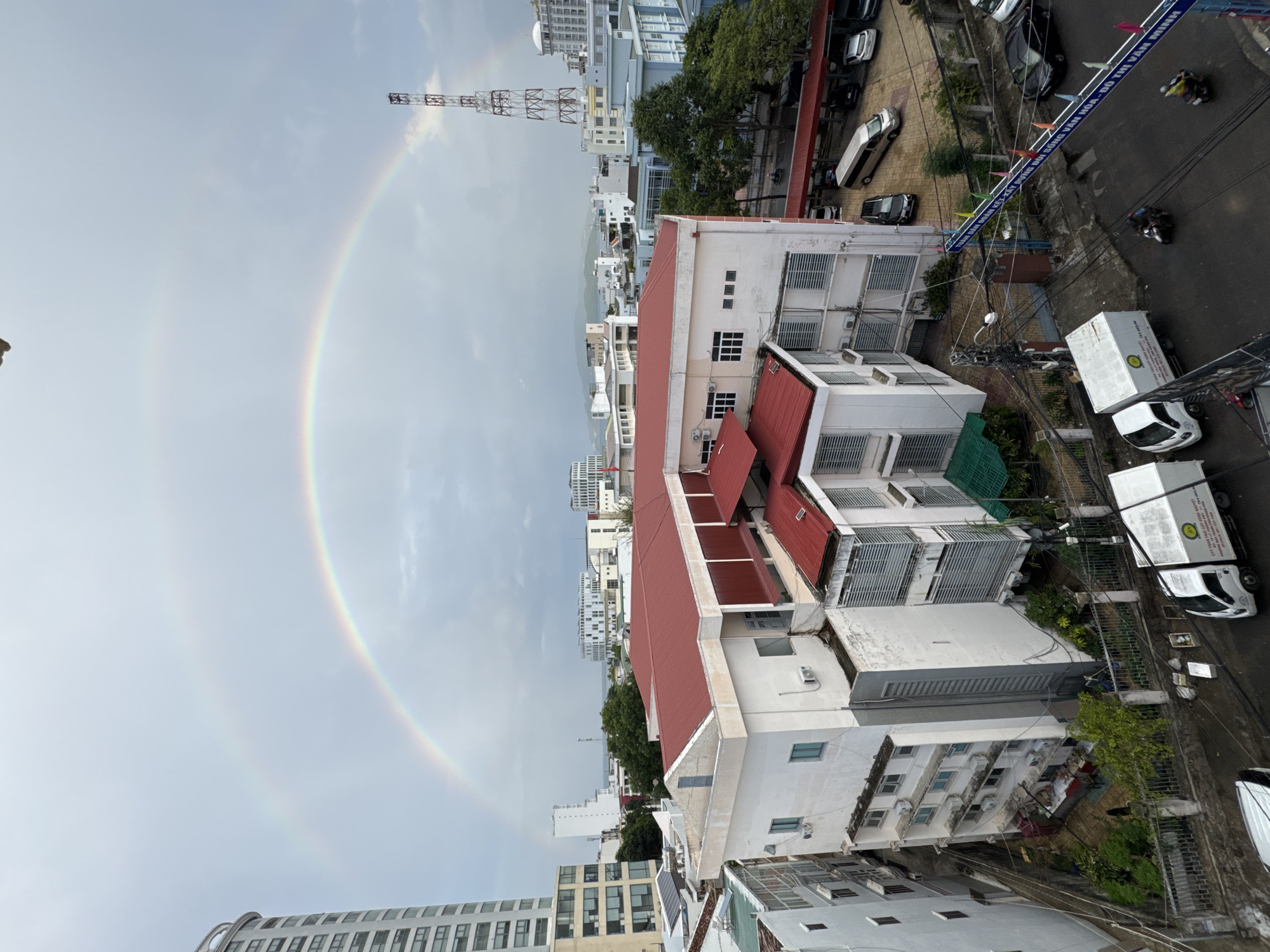 The image depicts a double rainbow arching over the city of Nha Trang, Vietnam, on an overcast day. The foreground features a white, multi-story building with a red tiled roof. To the left, a tall, modern skyscraper is visible. In the background, various other buildings of different architectural styles are scattered across the cityscape, leading towards the sea. A telecommunications tower stands prominently to the right of the frame. The street level shows parked cars, including vans and sedans, and a few motorcycles with riders. The overall scene suggests a typical urban environment during or after a rainfall, as indicated by the presence of the rainbow and the wet appearance of the road surface.