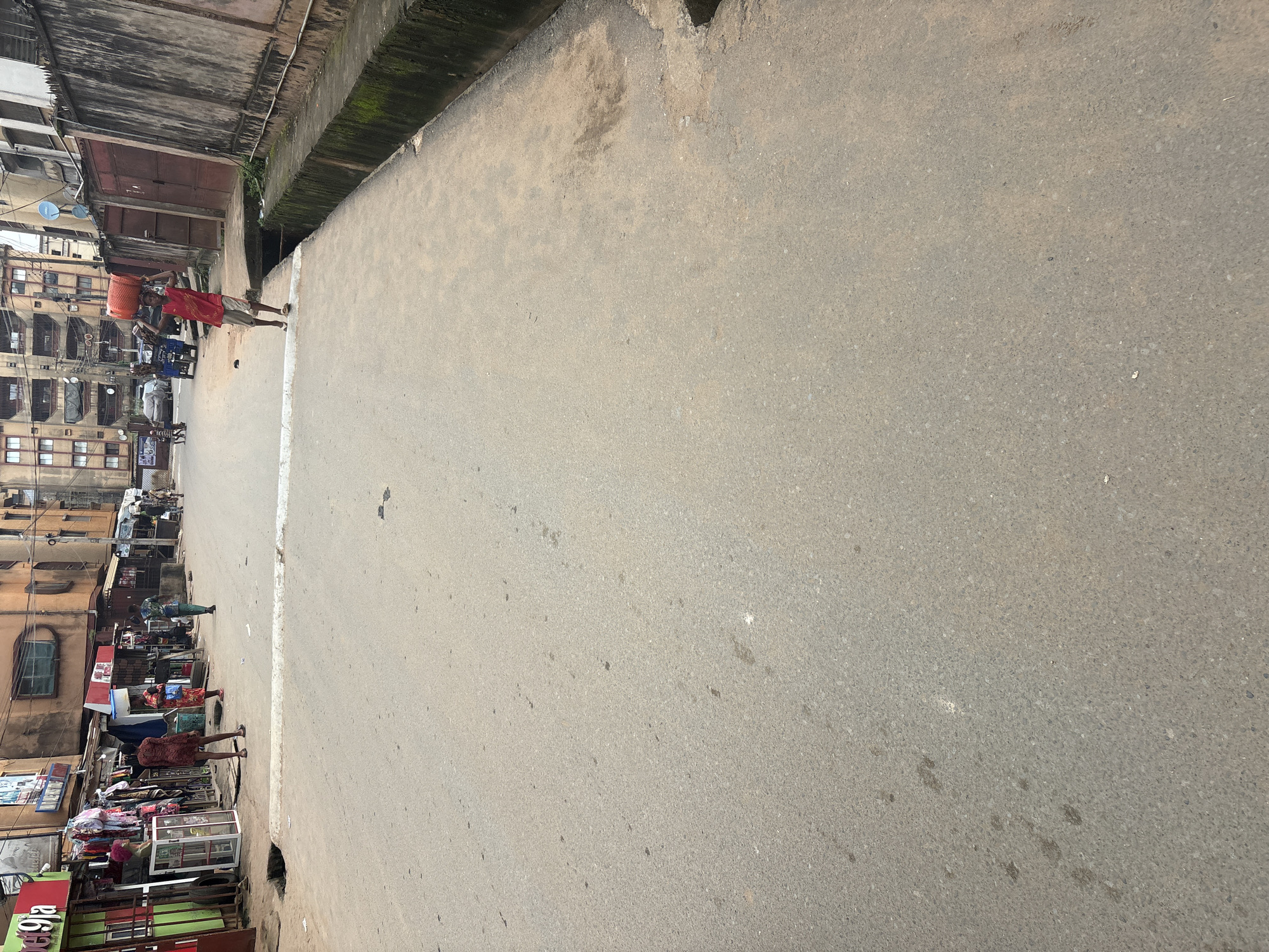 The image depicts a street scene in Onitsha, Nigeria. A young person in a red sleeveless shirt and beige shorts carries a large, orange basket on their head, walking on a raised concrete walkway next to a paved road. The road is worn and appears to be a main thoroughfare, with some litter scattered on its surface. On the left side of the street, there are storefronts with awnings and displayed goods, including clothing and mannequins. Several other individuals are visible in the background, walking and engaging in activities typical of a busy market street. Buildings line the opposite side of the street, some appearing aged and multi-storied. A drainage ditch runs alongside the walkway, filled with some debris and green growth. The overall environment suggests a lively urban commercial area.