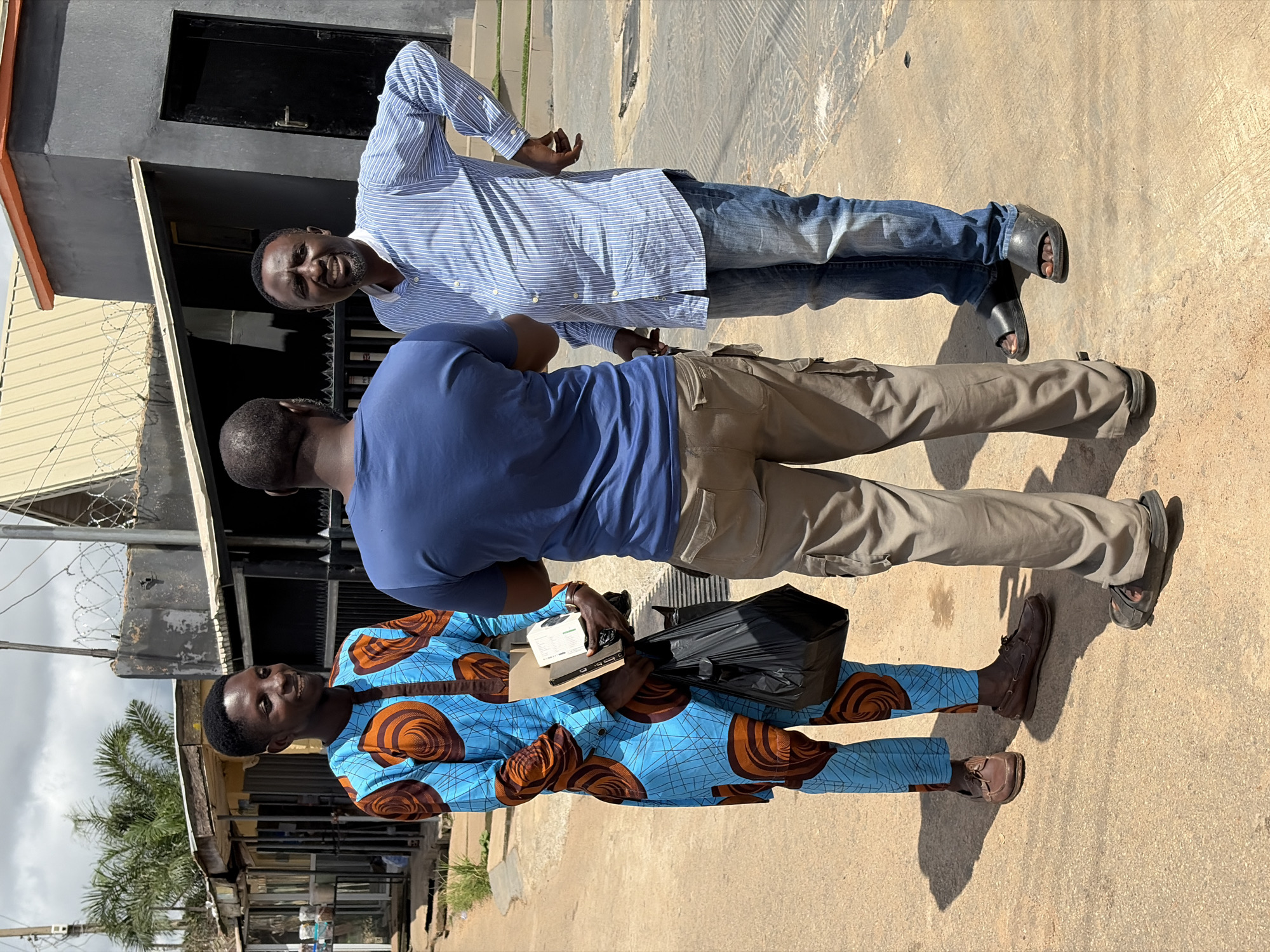 Three men are standing on a paved street in an urban area that appears to be Jos, Nigeria. The man on the left is wearing a blue and orange patterned traditional African outfit and brown loafers. He is holding a black plastic bag and a small box in his left hand, and he is smiling. The man in the middle is wearing a blue t-shirt and khaki cargo pants, with his back to the camera. The man on the right is wearing a blue and white striped button-down shirt and blue jeans. He is looking at the camera and has his hands on his hips. In the background, there is a building with a dark grey facade and a black door, along with some shops and barbed wire fencing. The sky is cloudy.