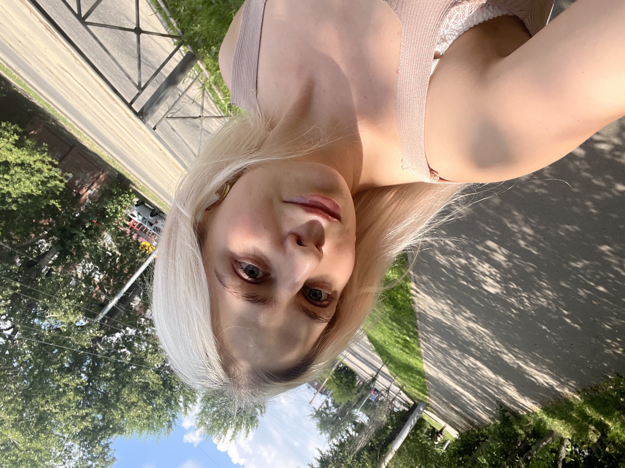 A young woman with blonde hair takes a selfie outdoors in Irkutsk, Russia. She is wearing a light pink top and a white lace bra. The background shows a sidewalk, a road, trees, and a blue sky. The lighting suggests it is daytime and sunny.