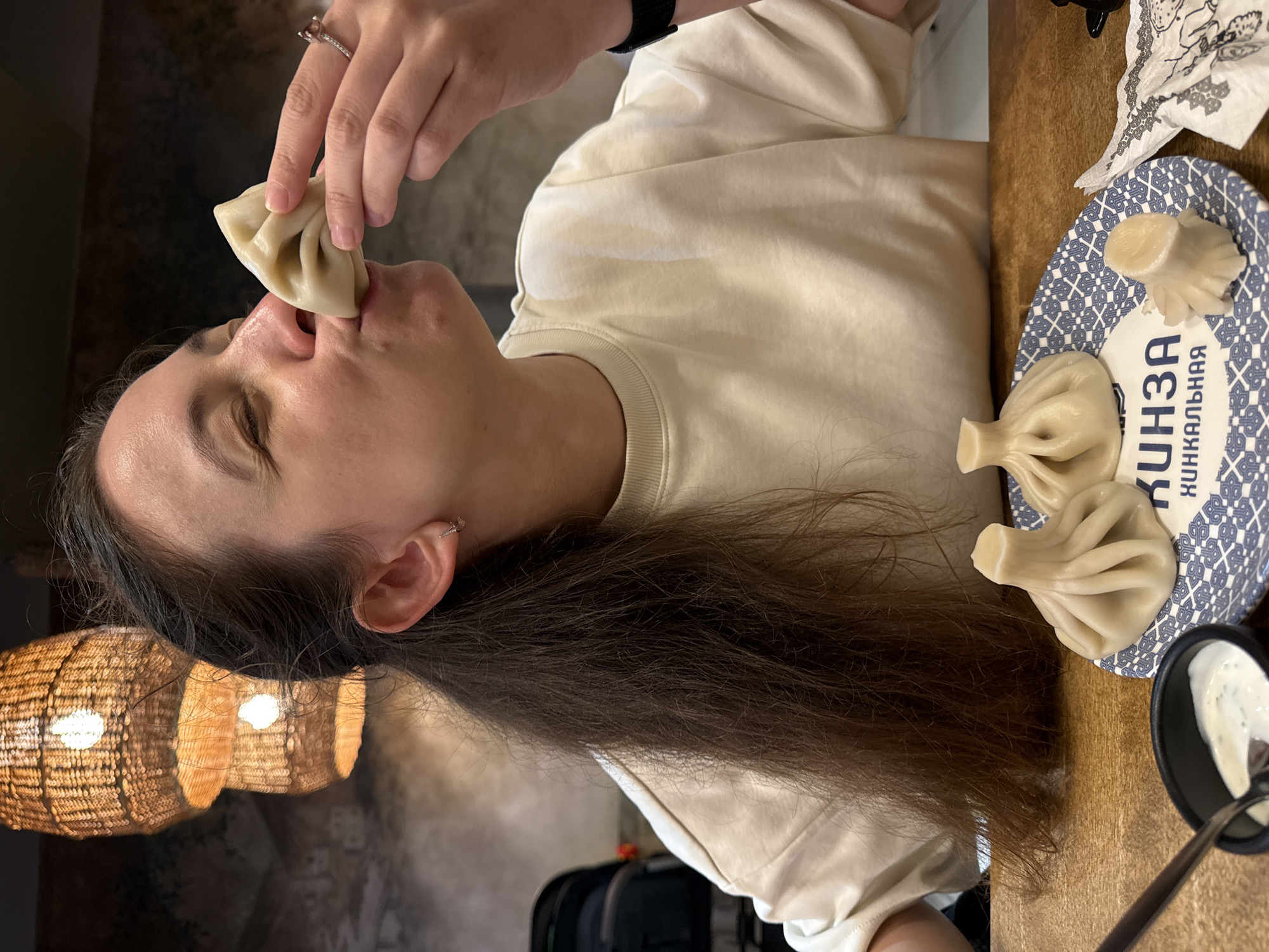 A woman is shown indoors, about to eat a khinkali, a type of dumpling. She is holding the khinkali to her nose and mouth with her right hand, her eyes closed in anticipation. She is wearing a light-colored crew neck sweatshirt and a ring on her right ring finger. Her dark brown hair is long and flowing. In the foreground, on a plate with a blue and white pattern, are two other khinkali and a portion of a third. To the left, a small black bowl contains a white sauce with herbs and a spoon. The background features two woven lamps hanging from the ceiling, providing warm lighting. The setting appears to be a restaurant. The text on the plate is in Cyrillic, reading "КИНЗА" and "ХИНКАЛЬНАЯ". This suggests the restaurant is named "Kinza" and specializes in khinkali. While the image itself does not provide specific geolocation data for Izhevsk, Russia, the presence of Cyrillic script is consistent with a Russian location.