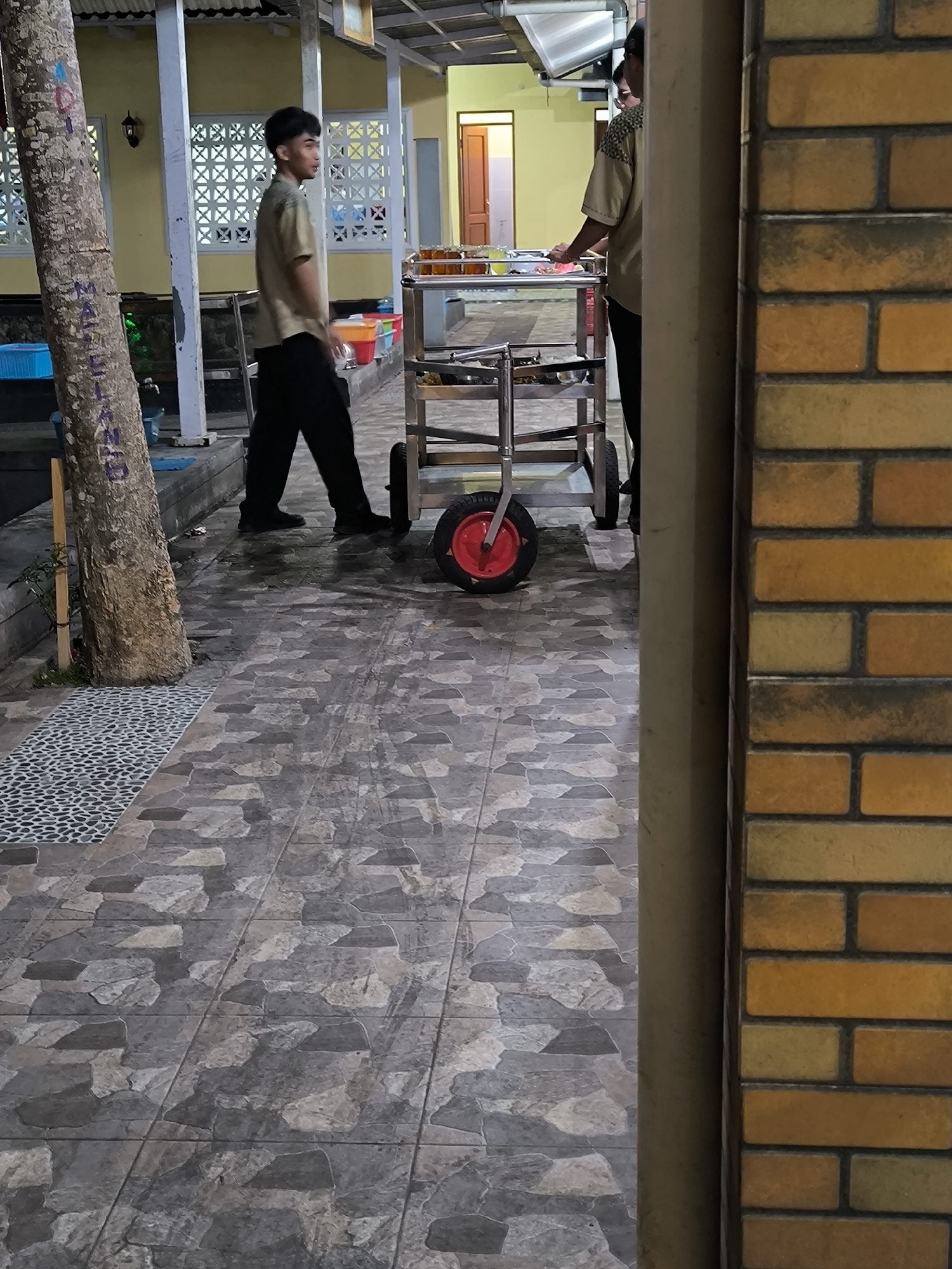 A young man in a light brown uniform stands on a tiled walkway next to a tree. He is looking to his left. Behind him, another person in a similar uniform is standing next to a food cart. The food cart has several metal trays filled with food. To the left of the walkway, there are white posts and a yellow wall with decorative windows. The walkway has a pattern of large, irregular-shaped tiles in shades of gray and brown. A brick wall with yellow and orange bricks is visible on the right side of the image. The lighting suggests it is either early morning or late evening.
