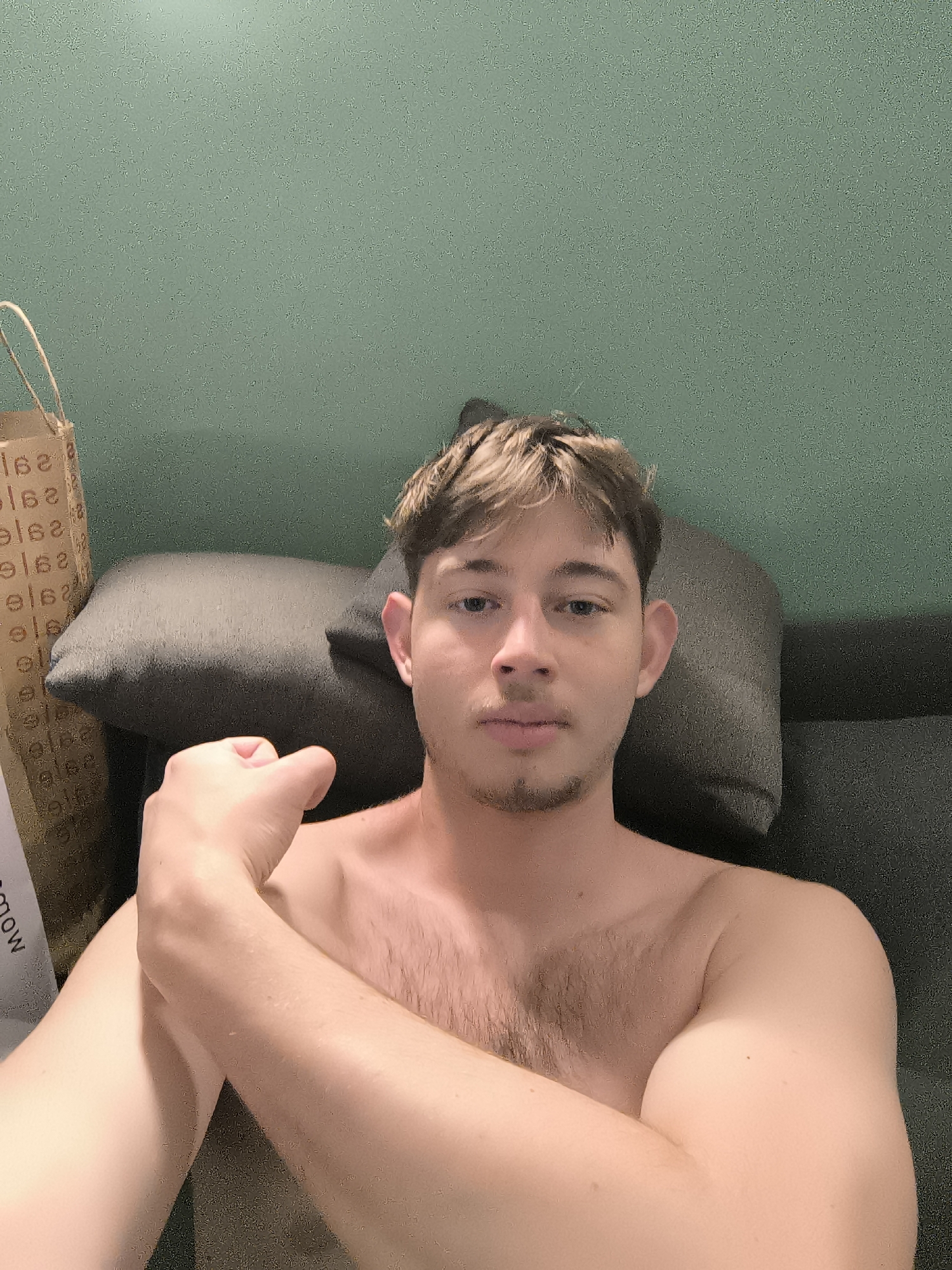 A chest-up shot of a shirtless fair-skinned young man with brown hair and stubble, possibly sitting on a sofa. He's looking directly at the camera, with his right arm bent and hand clenched into a fist positioned near his shoulder. Behind him is a gray pillow and a green wall. A paper shopping bag with text that appears to say "Sadow" is visible on the left. Location details are not provided in the image and thus cannot be determined.