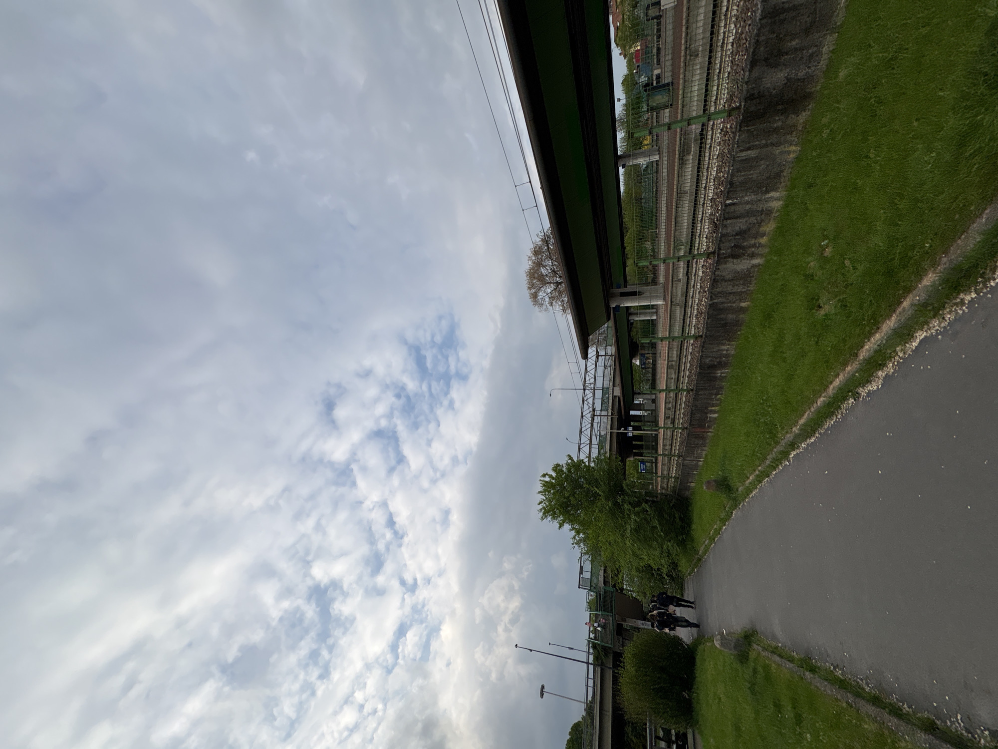 The image depicts a sideways view of a path running alongside a railway line in Milan, Italy. The sky is overcast with puffy clouds. To the right of the path is a grass verge and a concrete wall that supports the railway line. A railway bridge spans above, with electrical cables visible. A tree grows close to the bridge. On the path, several people are walking away from the viewer. Streetlights are present near the path. The railway tracks and some infrastructure can be seen behind a fence.