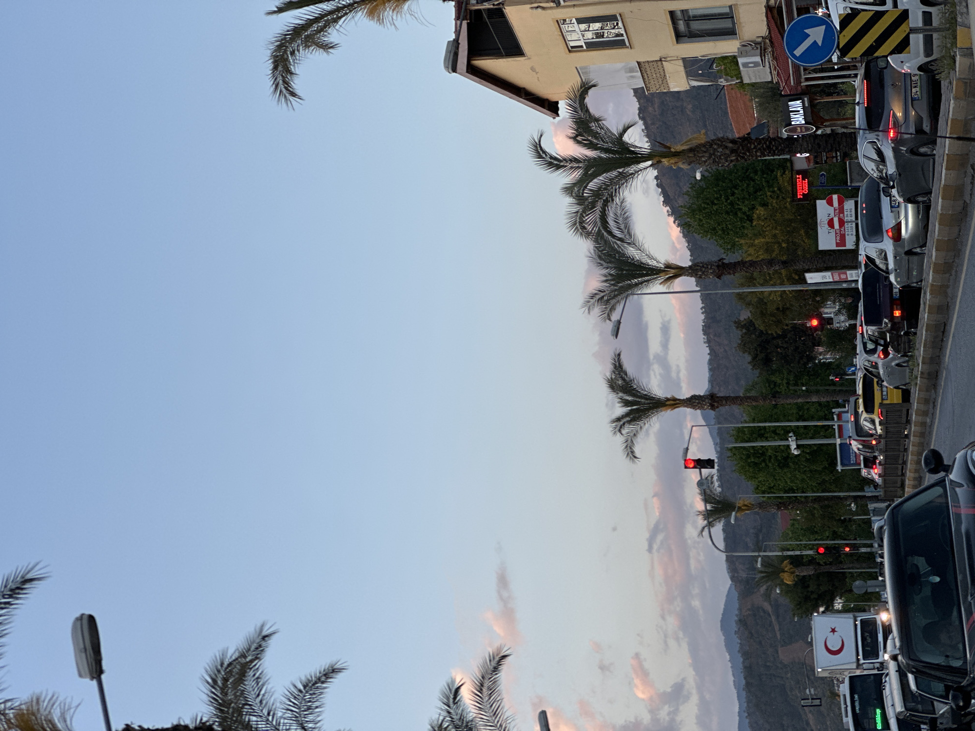 The image shows a street scene in Fethiye, Turkey. The sky is light blue with some clouds. Palm trees line the street, and cars are parked in a long line. A traffic light is visible, with a red light illuminated. The sign "BAKLAVA" is present, indicating a shop selling Turkish desserts. Other signs with Turkish text are visible, including "FISTIKLI ÖZEL" and "PROJE ATILI". A white vehicle with the Turkish flag is also seen. The background includes mountains and buildings.