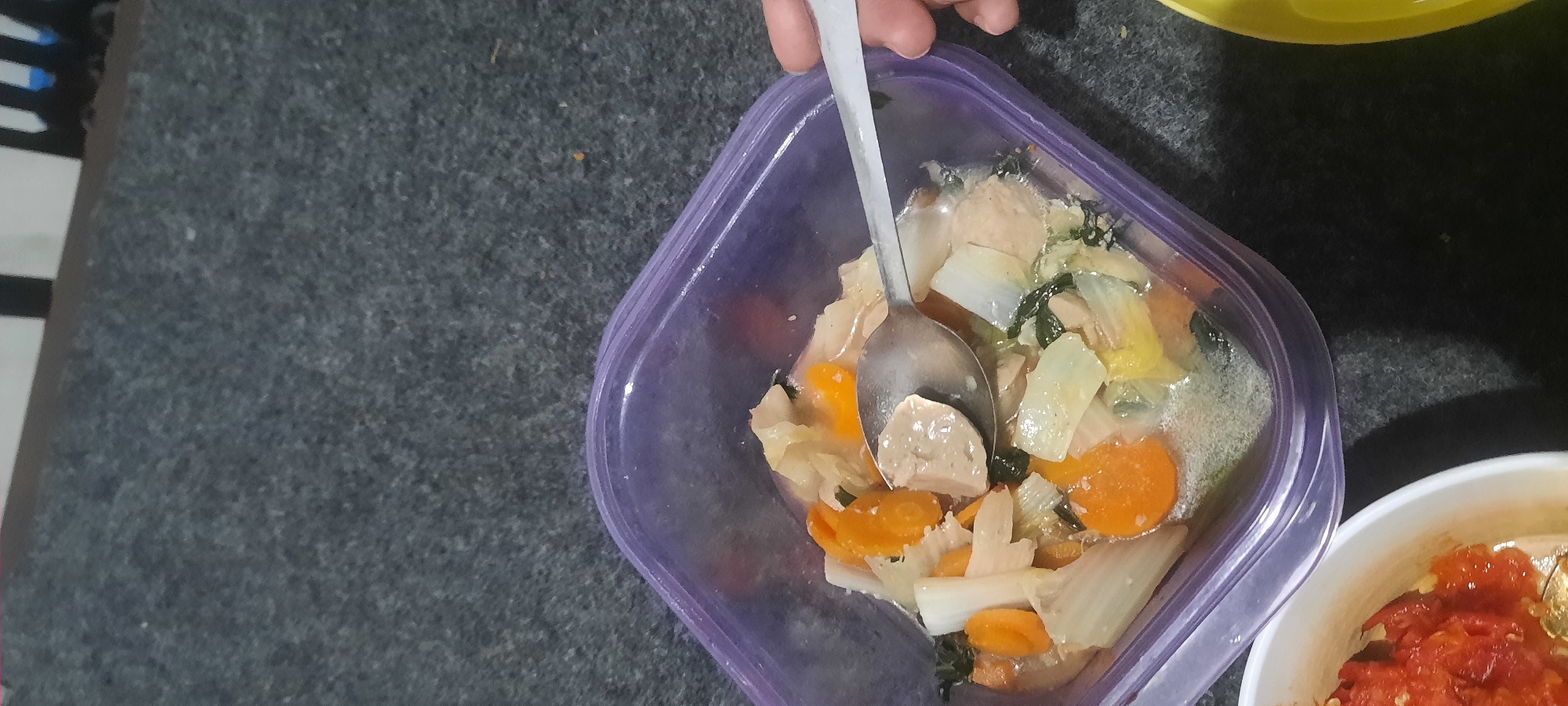 A close-up, overhead shot shows a purple plastic container filled with a vegetable and meat stew. The stew contains sliced carrots, bok choy, and meatballs in a light broth. A silver spoon, held by a person's hand which is partially visible on the right side of the image, is scooping up a meatball from the stew. The container is placed on a dark gray, textured surface that appears to be a carpet. To the left of the container, a white bowl containing a red, chunky sauce, possibly sambal, is visible. The lighting suggests an indoor setting, possibly a home kitchen in North Jakarta, Indonesia.