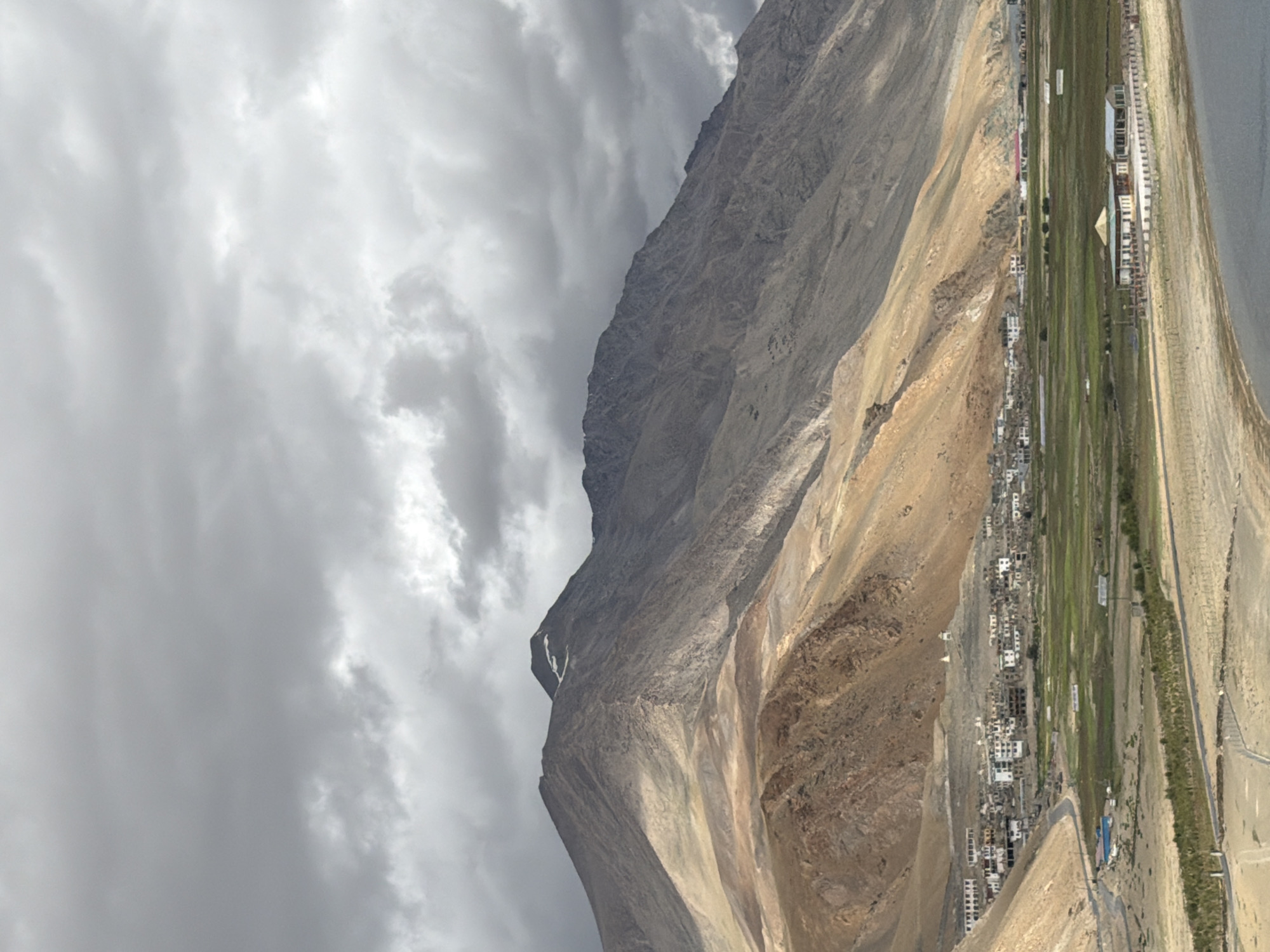 The image depicts a village scene in Karzok, India, nestled in a valley at the foot of a large, rugged mountain range. The sky is overcast with dramatic, dark clouds, suggesting a recent or impending storm. The mountains exhibit layers of brown and tan rock, with some patches of lighter scree and sparse vegetation.
In the midground, a cluster of white buildings, forming the village of Karzok, is situated on a gentle slope. These structures appear to be low-rise, with many flat roofs. A road winds through the village and extends outwards. Adjacent to the village is a vibrant green, flat area that looks like cultivated fields or a meadow.
In the foreground, a body of water, likely a lake, is visible, with its surface reflecting the muted light. Along the edge of the water and extending towards the village are areas of dry, sandy terrain. A low fence-like structure runs along the embankment between the water and the land. Further inland, there are more arid, sandy areas with sparse vegetation and winding paths or roads.
A distinct building with a light-colored roof and a surrounding courtyard is positioned prominently on the edge of the dry land, closer to the lake. The overall impression is one of a remote, high-altitude settlement with a stark, natural environment. There are no visible people or animals in the image.