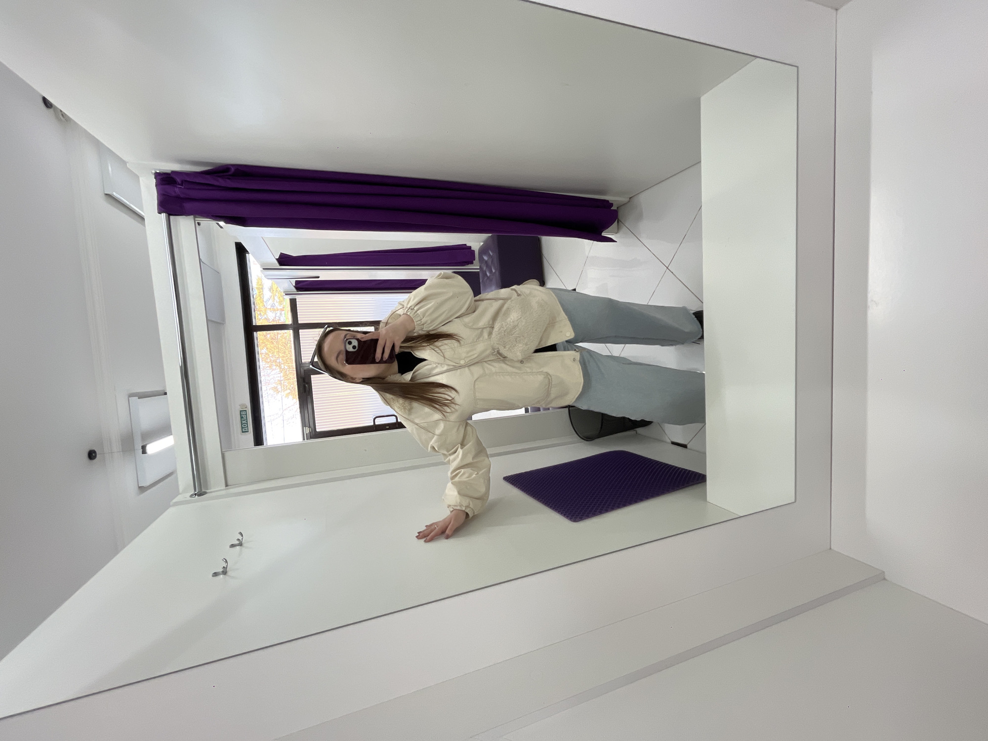 A person is visible in a mirror located in a dressing room in what appears to be a store in Novopokrovskaya, Russia. The individual is holding a smartphone and taking a selfie. The dressing room is characterized by white walls and tiled flooring, with a purple curtain and a dark purple floor mat present. A window is visible behind the person. Clothing hooks are visible on the wall.