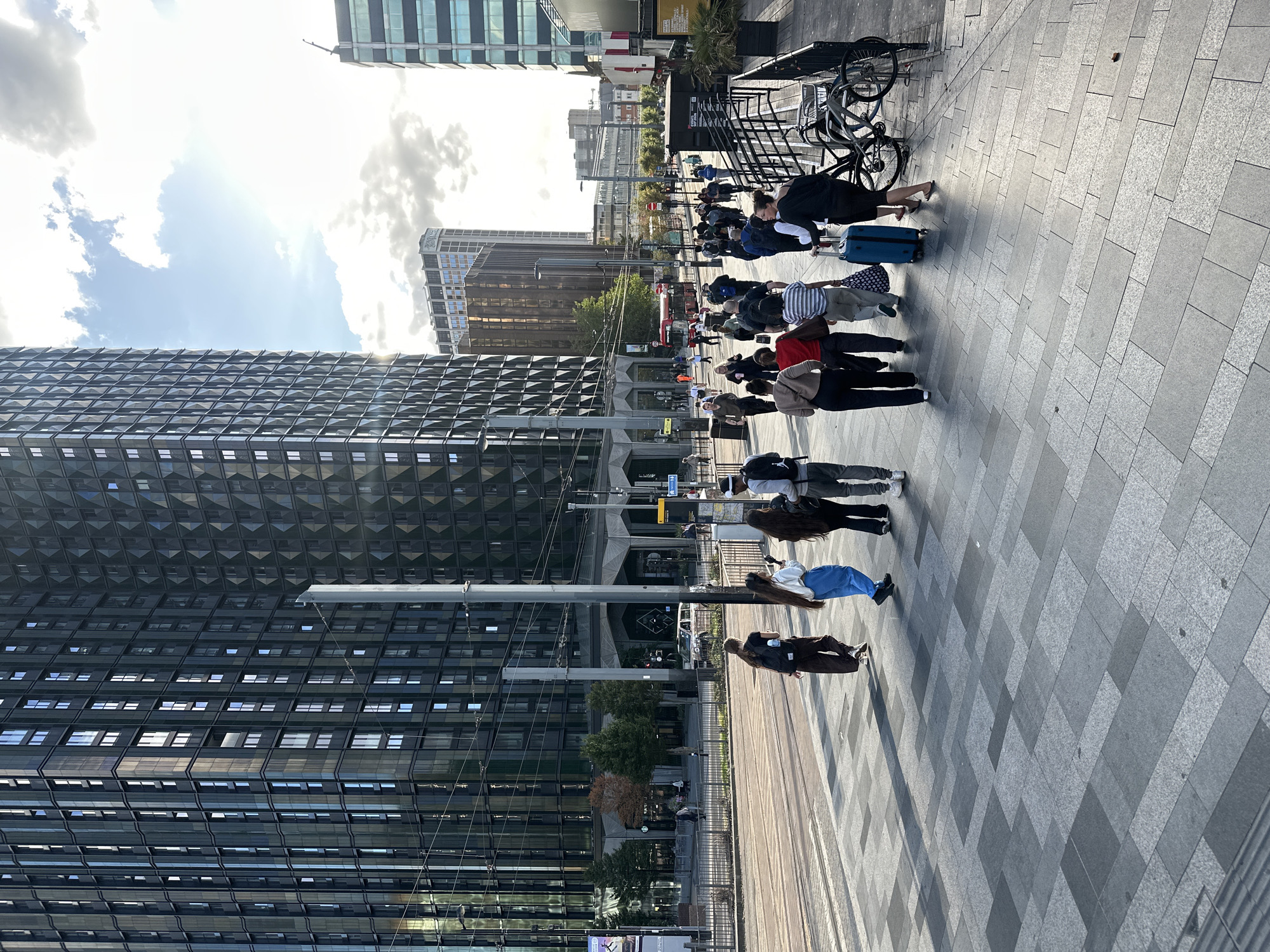 In the city of Unknown, Unknown, a street-level view captures a bustling scene with a focus on a pedestrian walkway and the surrounding urban environment. The foreground features a paved area with large, grey, rectangular tiles arranged in a staggered pattern, with lighter and darker shades creating a textured appearance. Sunlight casts distinct shadows on the pavement, indicating a bright day.
A group of people are walking away from the camera, mostly towards the right side of the frame. They are dressed in casual attire, with some carrying bags and one person pulling a blue suitcase. Their movement suggests they are commuting or traveling.
In the mid-ground, a street with tram tracks is visible, with no trams currently present. A few trees with autumn foliage are planted alongside the street. Several buildings form the backdrop, most notably a very tall, modern skyscraper with a dark, reflective facade composed of numerous windows arranged in a grid pattern. Another building to its right has a similar dark facade, but with a diamond-shaped pattern. Further in the distance, other urban buildings are visible, including a lighter-colored tower.
A few other individuals and a white car are present in the mid-ground and background, indicating ongoing activity. Overhead, utility poles and wires crisscross the sky, typical of an urban setting. The sky is partly cloudy with patches of blue, and the sun is visible, contributing to the lighting and shadows. The overall impression is one of a modern, active city street on a sunny day.