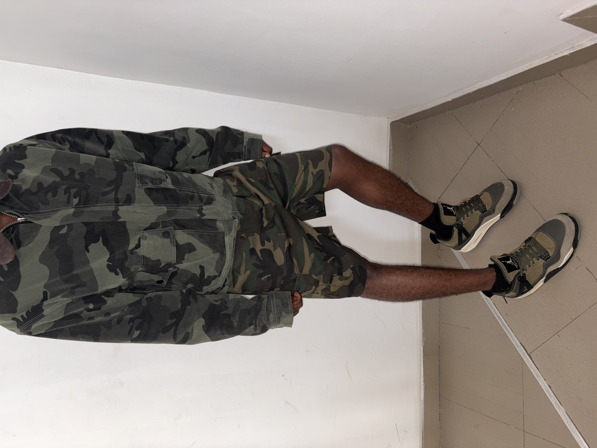 A person is standing in the corner of a room with light beige tile flooring and white walls. The person is wearing a matching camouflage jacket and shorts, black socks, and two-toned olive and gray athletic shoes. The location is in the city of Igboefon, Nigeria.