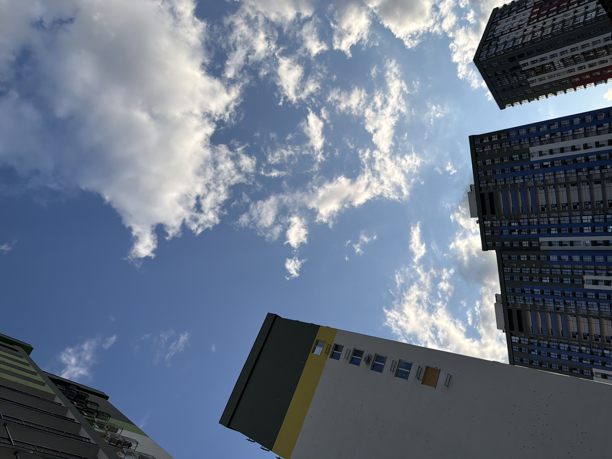 The image captures a low-angle view of apartment buildings against a backdrop of a blue sky with scattered white clouds. In the foreground, on the left, is a tall, light-colored building with a dark green and yellow band across its top. The building has several windows and air conditioning units visible. To the right and slightly behind this building, two other taller apartment buildings are visible. The one in the center has a facade primarily in shades of blue and grey, with numerous windows. The building on the far right has a more varied facade with sections of red, white, and grey. The perspective suggests these buildings are part of a modern urban development, likely in the city of Kyiv, Ukraine, given the architectural style. There are no people or vehicles visible in the frame, and the scene appears to be a static architectural study focused on the buildings and the sky.