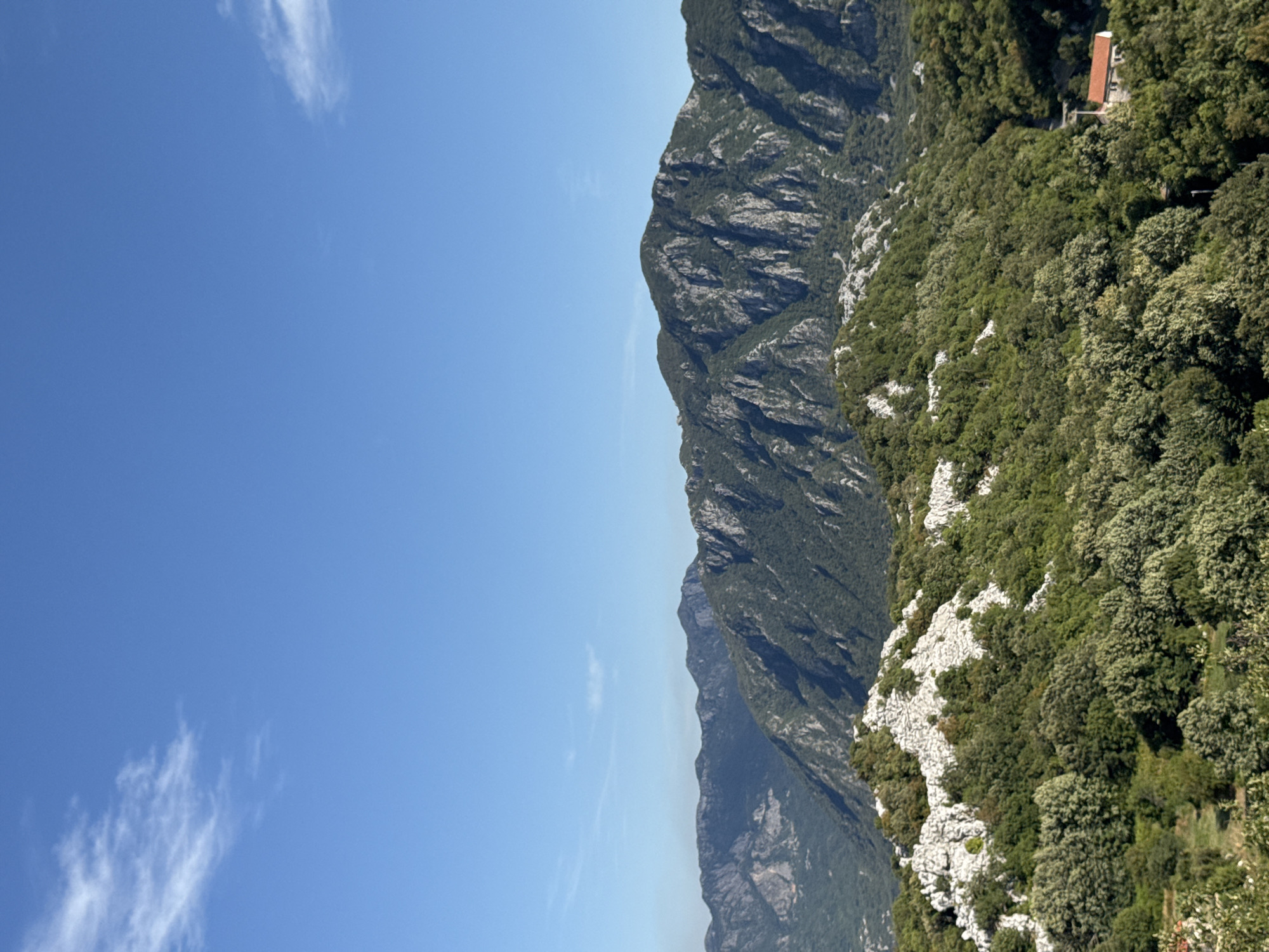 A panoramic, high-angle, eye-level shot captures a vibrant mountainous landscape under a clear, light blue sky with sparse, wispy white clouds. The foreground is dominated by lush green foliage and rocky outcrops. In the middle ground, the terrain rises into rugged, dark green mountains with stark, exposed rock faces. The background features a further range of hazy, bluish mountains stretching to the horizon. On the far right of the mid-ground, nestled among the trees, is a small building with a terracotta-tiled roof. The overall scene depicts a natural, undeveloped environment, likely in or around the village of Gluhi Do, Montenegro.