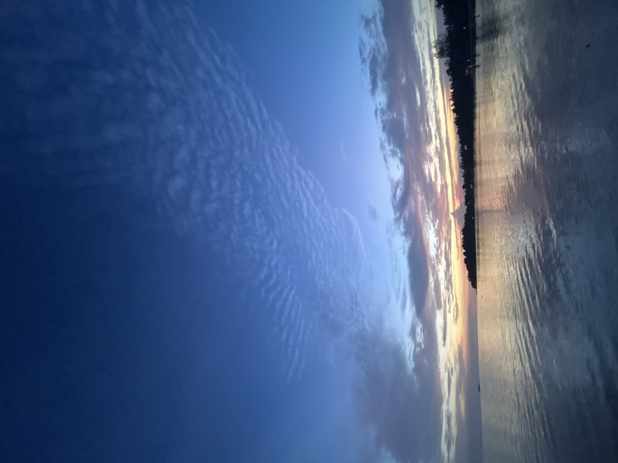 This image depicts a scenic view of a body of water, possibly a sea or large lake, at either dawn or dusk. The sky is a gradient of deep blue at the top, transitioning to lighter blues and then to warm hues of orange and pink near the horizon, indicating sunset or sunrise. Wispy clouds are scattered across the sky, some catching the last or first light of the sun.
On the right side of the image, the water reflects the colors of the sky, with ripples creating a textured surface. The left side of the image shows a silhouetted shoreline with trees. A thin line of light is visible along the horizon, suggesting a low sun position. Some small, distant lights can be seen along the shoreline, possibly indicating a town or buildings. The overall mood of the image is serene and peaceful, capturing the natural beauty of the twilight hours. No people or specific objects are clearly visible. The location context suggests Ko Pha-ngan District, Thailand.