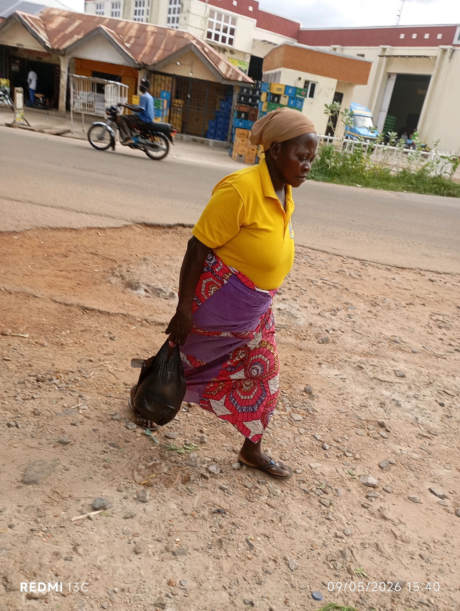 An elderly African woman walks on a dirt road in Shendam, Nigeria, carrying a black plastic bag in her right hand. She wears a yellow polo shirt and a purple and pink patterned wrap skirt, along with brown sandals. Her head is covered with a beige headscarf. In the background, a motorcycle rider in a blue shirt is visible on the paved road, passing by a row of shops. The buildings have corrugated metal roofs and display various goods, including crates of drinks. The scene depicts a typical street view in the city, with both paved and unpaved surfaces and commercial activity.