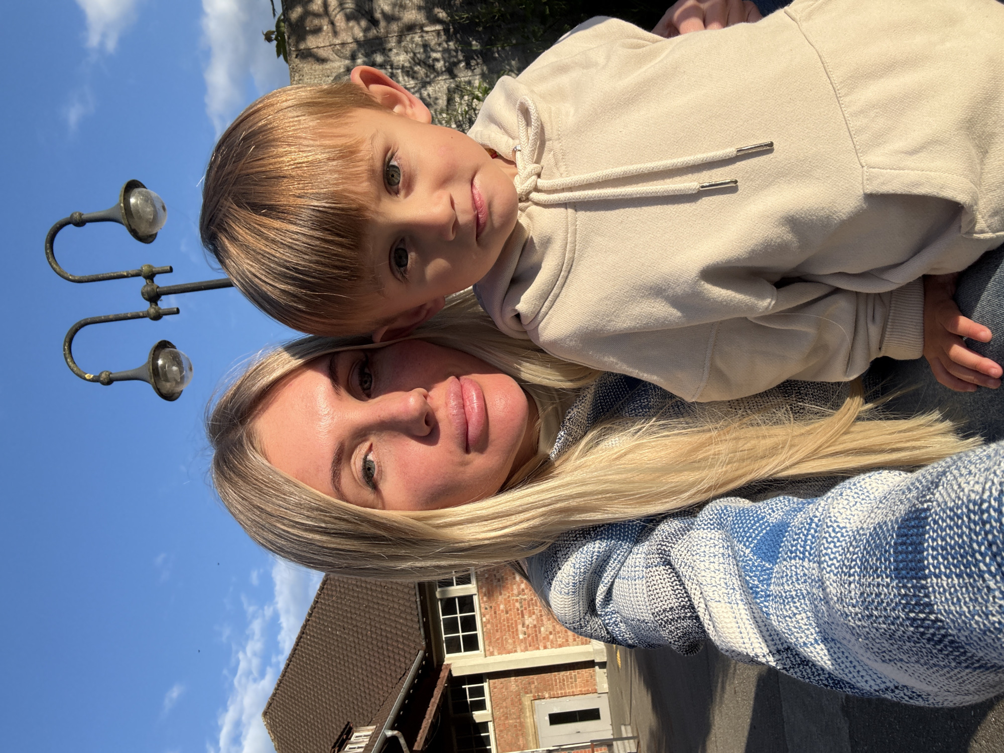 The image shows a selfie of a woman and a young child. The woman has long blonde hair and is wearing a blue and white patterned jacket. The child has short brown hair and is wearing a beige hoodie. They are standing outside, with a blue sky and some clouds in the background. In the background, there's a building made of brick with white-framed windows. Also in the background, there's a three-bulb lamppost. Based on visual cues and contextual knowledge, the location appears to be in the city of Riedlingen, Germany.