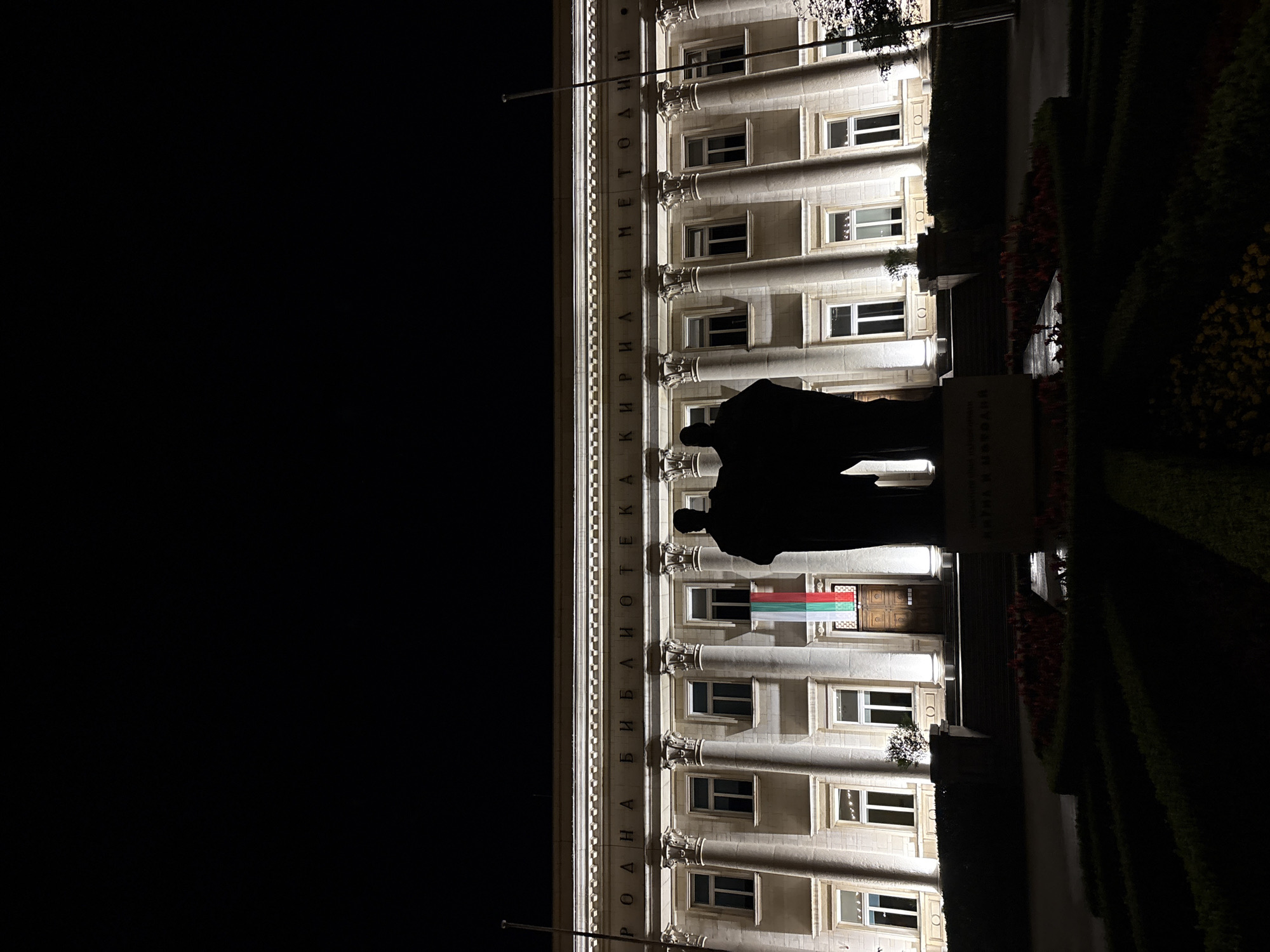 The image depicts the Sts. Cyril and Methodius National Library in Sofia, Bulgaria, at night, illuminated by artificial lighting. The grand building features a classical architectural style with multiple stories, numerous windows, and prominent columns along its facade. Running vertically along the upper section of the building's facade is the Cyrillic inscription "НАРОДНА БИБЛИОТЕКА КИРИЛ И МЕТОДИИ," which translates to "National Library Sts. Cyril and Methodius."
In front of the library, silhouetted against its lit facade, stands a large monument. The monument clearly shows the dark figures of two individuals, identified by the inscription on its base as "СВЕТИ КИРИЛ И МЕТОДИИ" (Saints Cyril and Methodius). A Bulgarian flag, displaying its characteristic white, green, and red horizontal stripes, hangs from a window or balcony on one of the lower levels of the building.
The scene is captured at night, with a completely dark sky, emphasizing the warm glow of the building's illumination. The foreground features dark, manicured hedges and some hints of floral arrangements, suggesting a landscaped area or plaza. No specific activity or event is taking place; the image presents a serene and majestic nighttime view of this significant cultural landmark in Sofia.