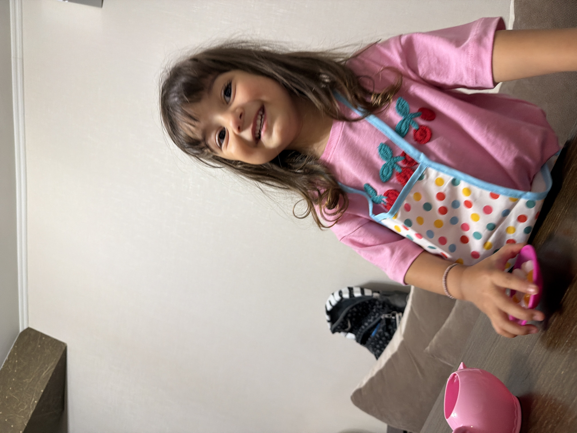 A young girl with dark hair and brown eyes, wearing a pink t-shirt with a colorful polka-dot apron featuring cherry embellishments, smiles at the camera. Her right hand, adorned with a thin bracelet, is reaching for a small pink dish containing several small, colorful candies. To her left, a pink teapot sits on a dark wooden surface. In the background, a beige couch and a backpack with a black and white striped pattern are visible against a plain, light-colored wall. The scene appears to be indoors, likely in a home environment. The image does not provide any information about the location in Burgas, Bulgaria.