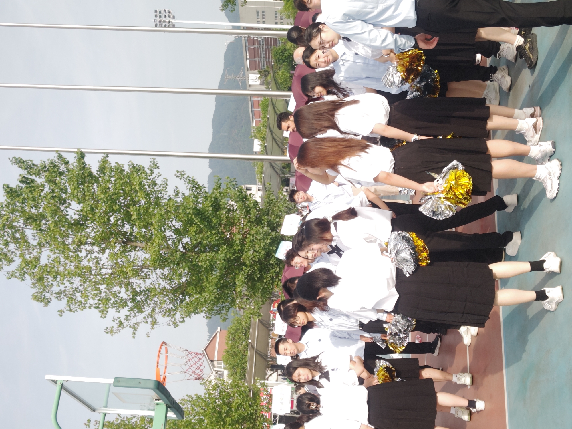 A group of students dressed in white shirts and black skirts is standing on an outdoor basketball court, holding silver and gold pom-poms. The location appears to be in a school area, potentially in the Chongqing High-tech Zone in China. Trees and buildings are visible in the background, along with a mountain range. A basketball hoop is visible.