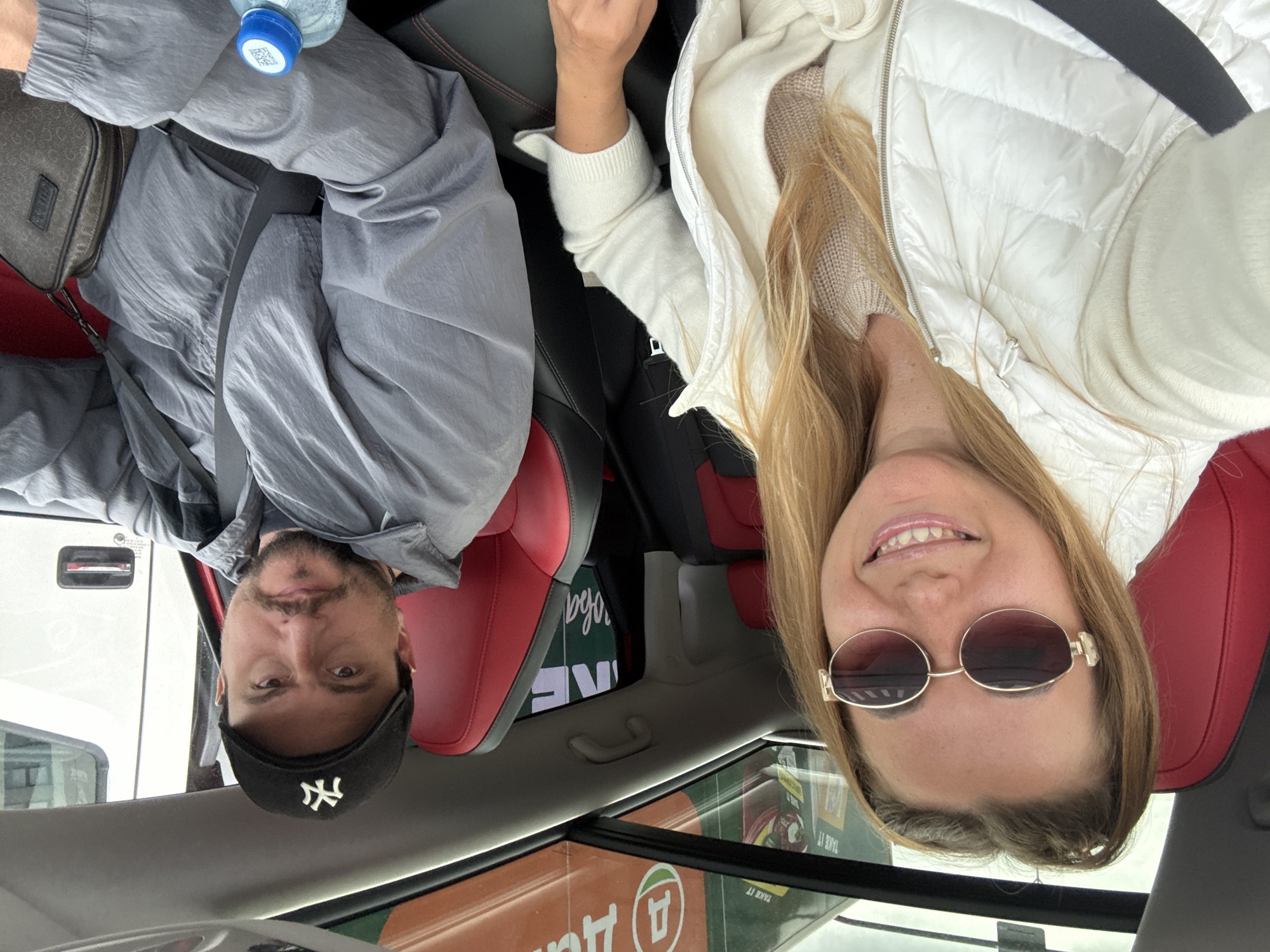 A woman and a man are sitting in the front seats of a car. The woman is on the left, wearing a white puffer vest over a cream-colored sweater and large oval sunglasses. She is smiling and has blonde hair pulled back. The man is on the right, wearing a black baseball cap with a white "NY" logo, a gray jacket, and a dark crossbody bag. He is looking forward with a neutral expression. The car has red and black interior seating and a panoramic sunroof. Outside the car, a sign with the letter "Д" in a circle and some Cyrillic text is visible through the windshield and sunroof. A bottle of water with a blue cap and a QR code is on the man's lap.