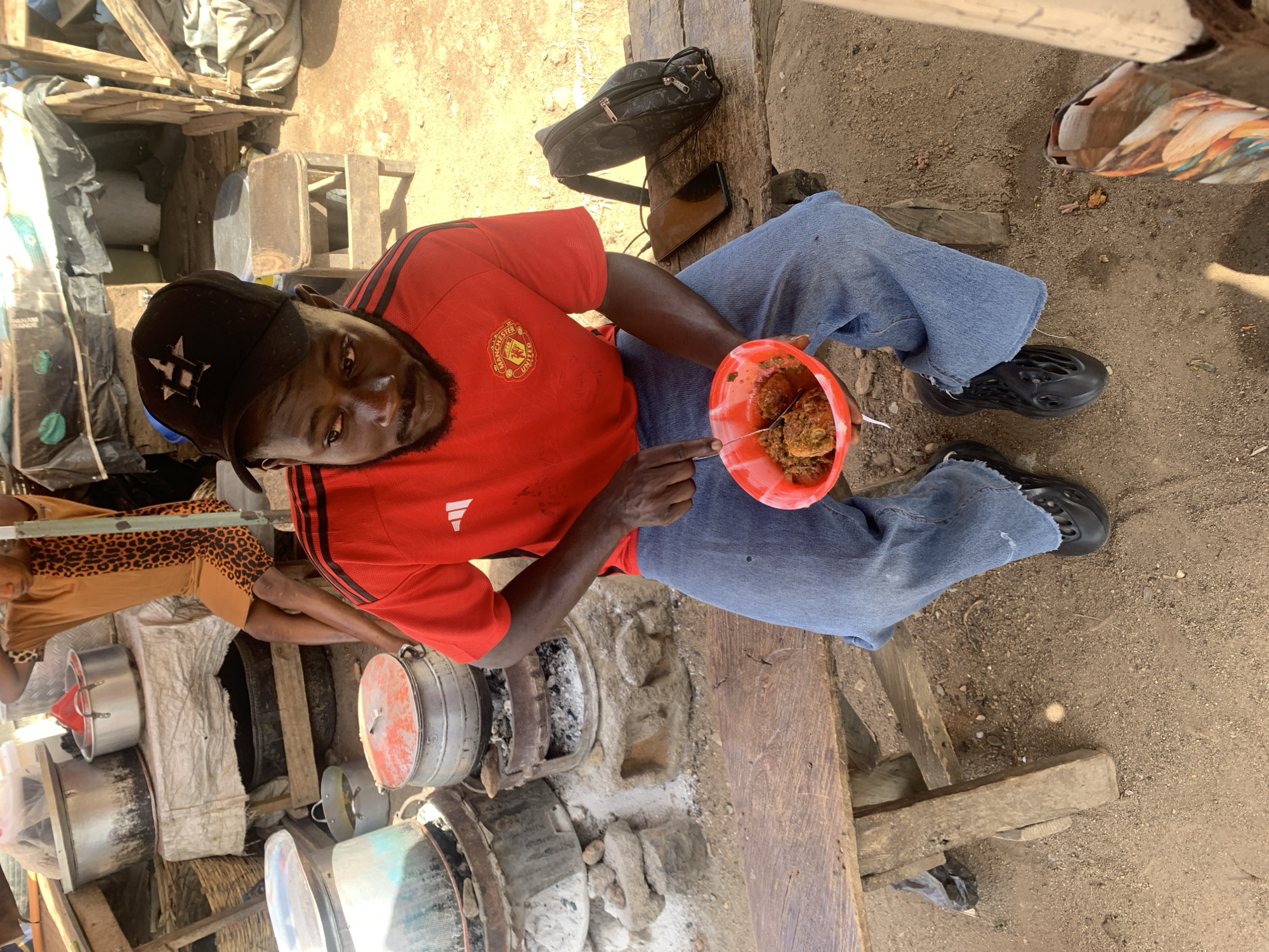An eye-level, overhead shot shows a man eating street food outside in Rafin Par, Nigeria. The man is seated on a wooden bench, wearing a red Manchester United t-shirt with black stripes on the shoulders, a black baseball cap, and blue jeans. He holds a red bowl filled with food, which he is about to eat with a utensil. His black shoes are visible on the ground.
Adjacent to him is a cooking area with several large metal pots on primitive stoves, and a woman wearing a leopard-print shirt is visible near these pots. Behind the man is a black bag and a mobile phone. The area appears to be a makeshift outdoor kitchen or food stall. The background shows a cluttered urban environment. The ground is dirt and gravel.